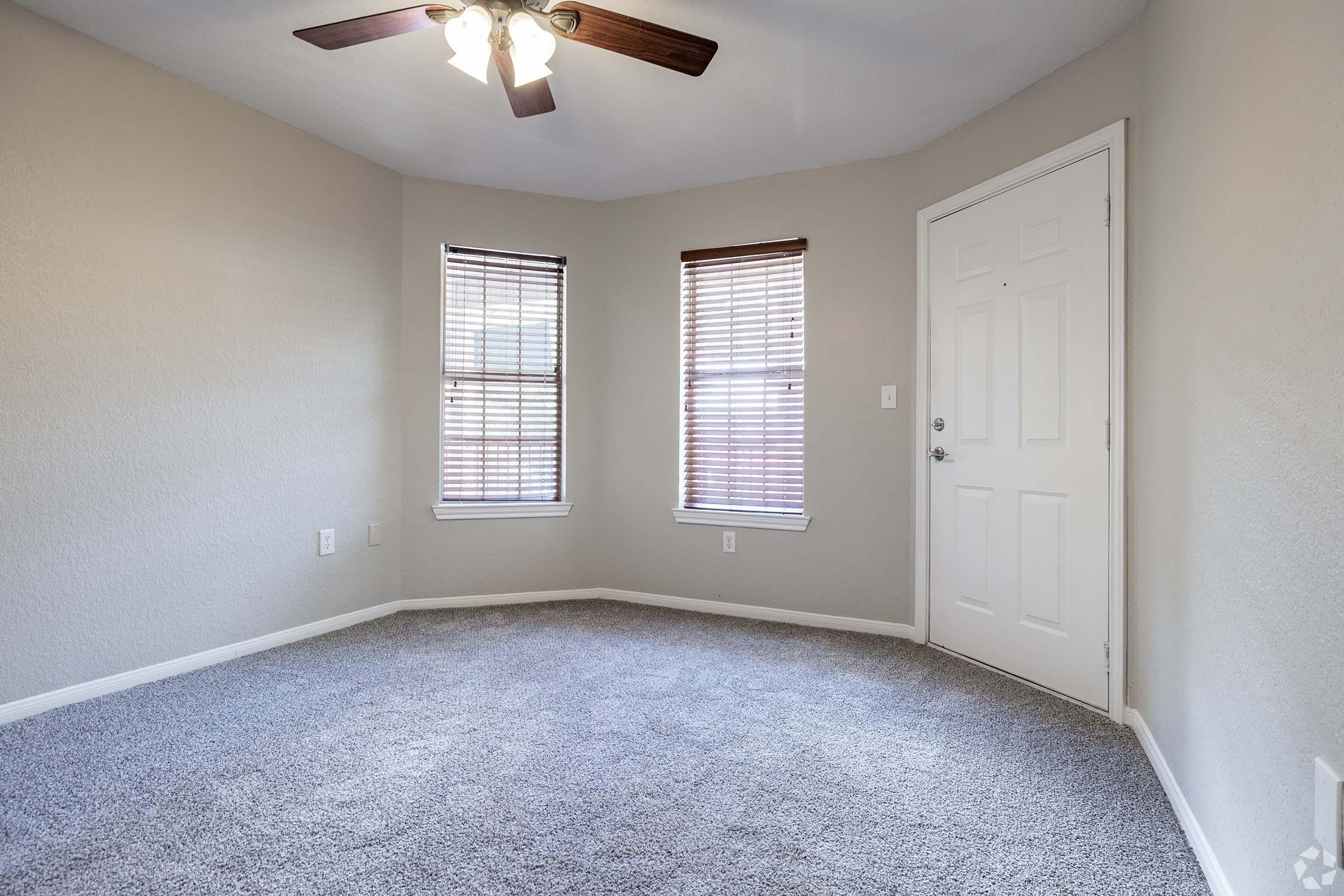 Empty room with light gray walls and carpet, featuring two windows with blinds and a ceiling fan. The door is white and located on the right side. The lighting is bright, creating a spacious and clean atmosphere.