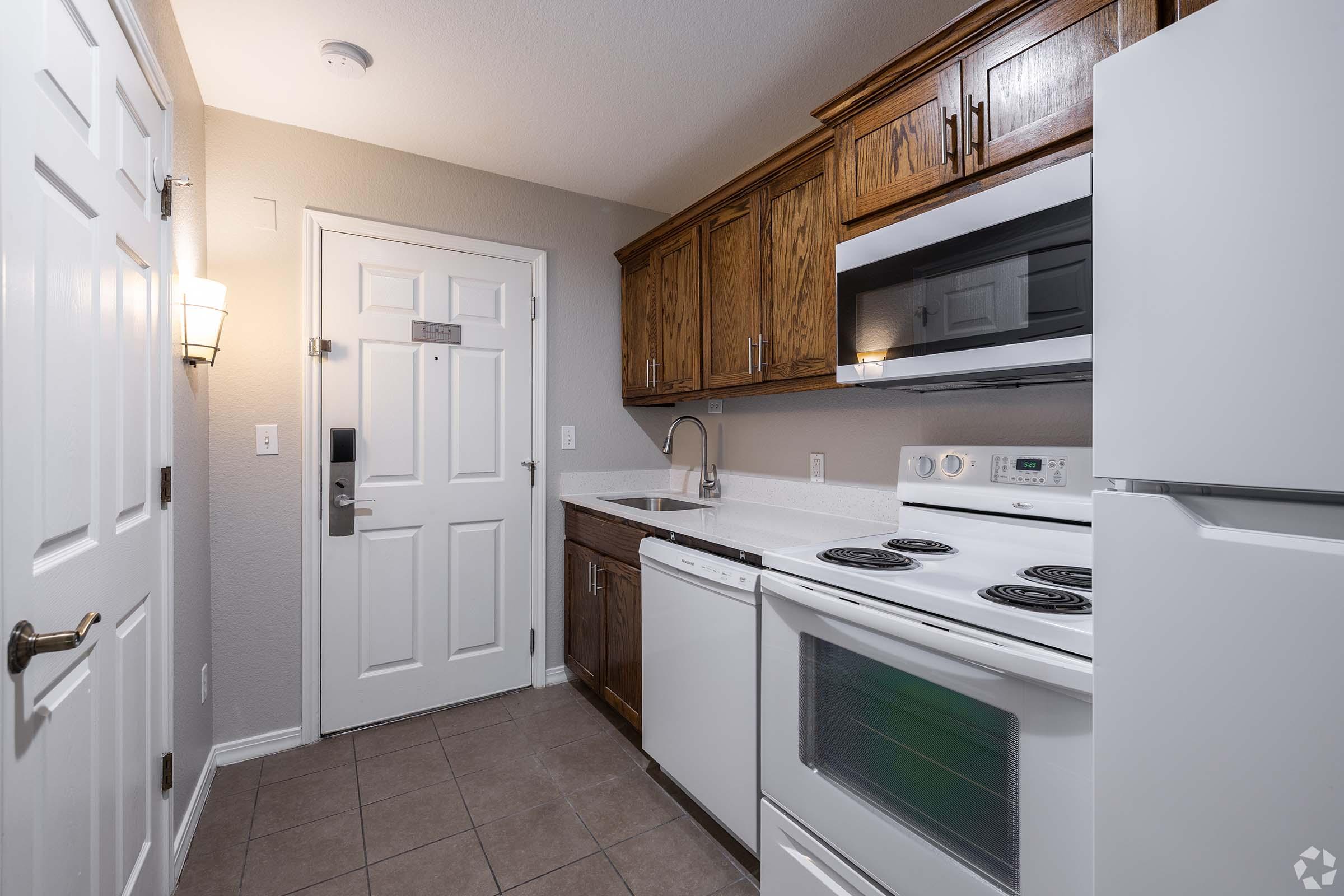 A well-lit kitchen featuring wooden cabinets, a white dishwasher, an electric stove, and a microwave. The entrance door is visible on the left, and the flooring is tiled. The overall decor is modern and functional, with a clean, organized appearance.
