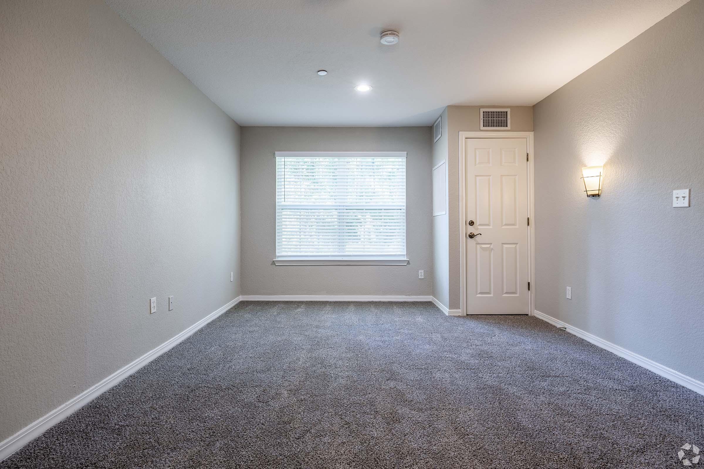 A spacious, well-lit room with grey walls and carpet. It features a window with blinds, a wall-mounted light fixture, and a door leading to another area. The overall atmosphere is clean and simple, suitable for various uses.