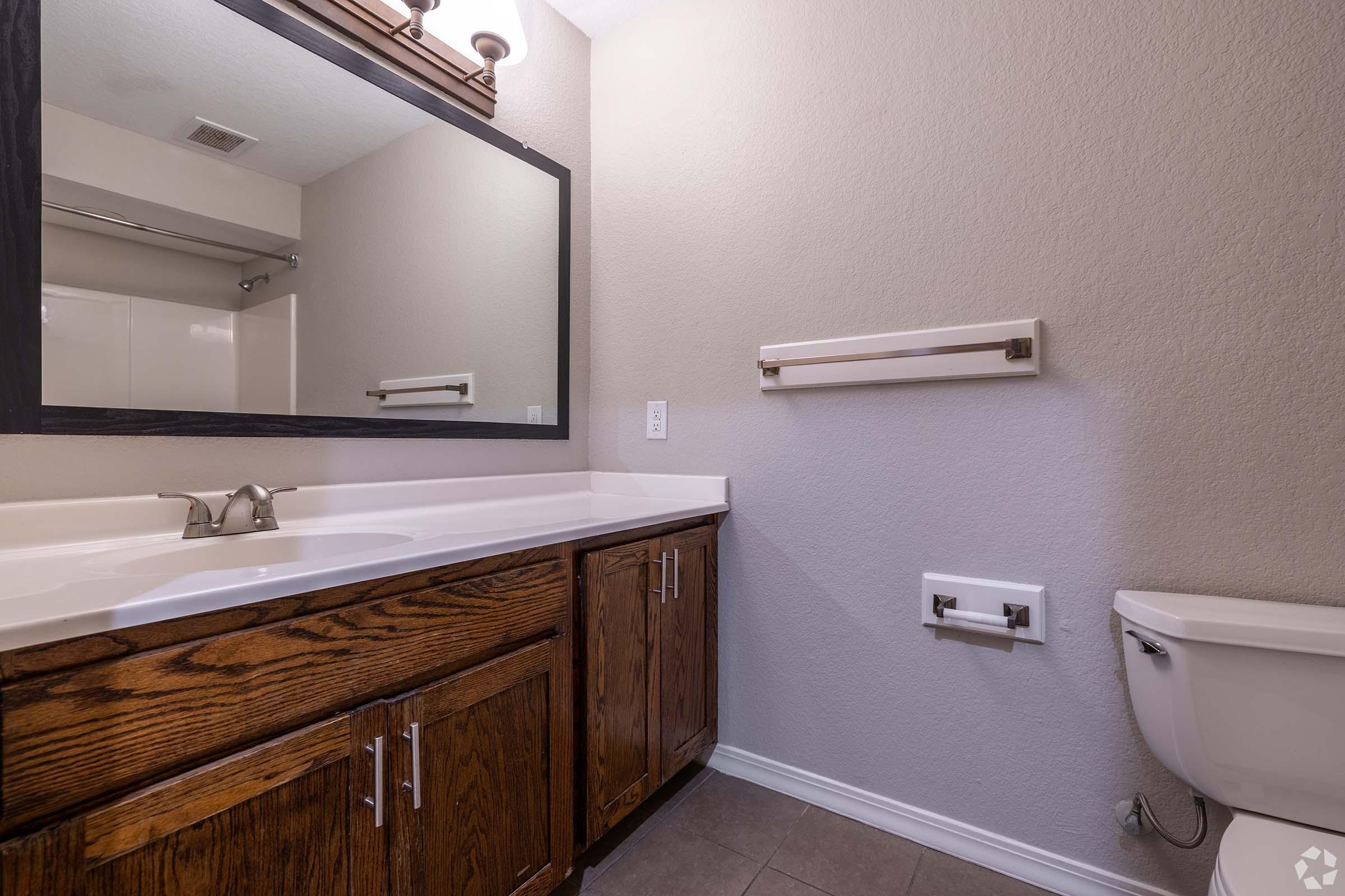 A clean and modern bathroom featuring a large mirror above a white countertop with wooden cabinets below. To the right, there's a toilet and a towel bar mounted on the wall. The walls are painted in a neutral tone, and the flooring is tiled, creating a spacious and well-lit appearance.