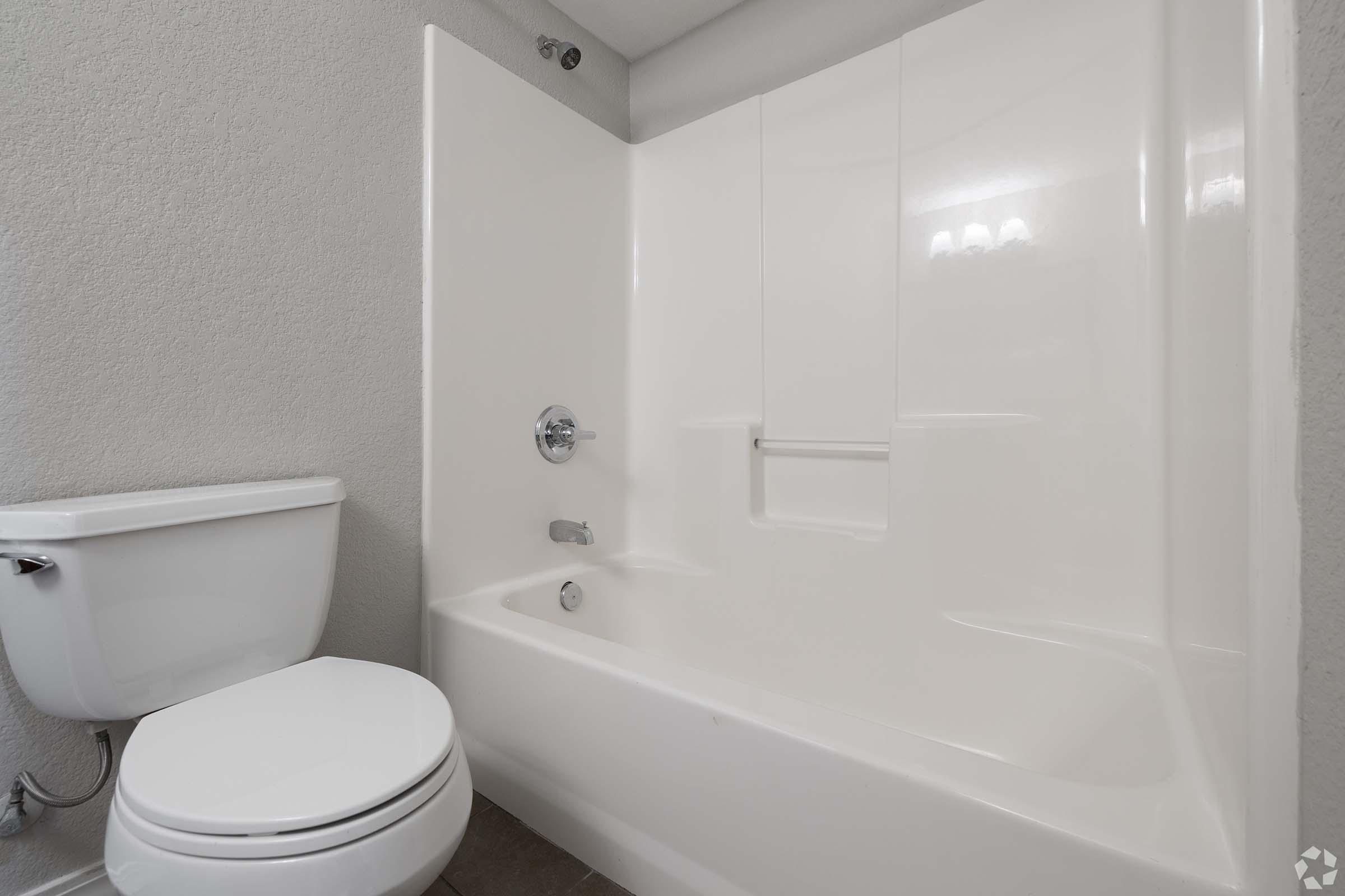 A clean bathroom featuring a white bathtub and shower combination, a modern toilet, and neutral-colored walls. The space is well-lit, showcasing a simple and contemporary design.