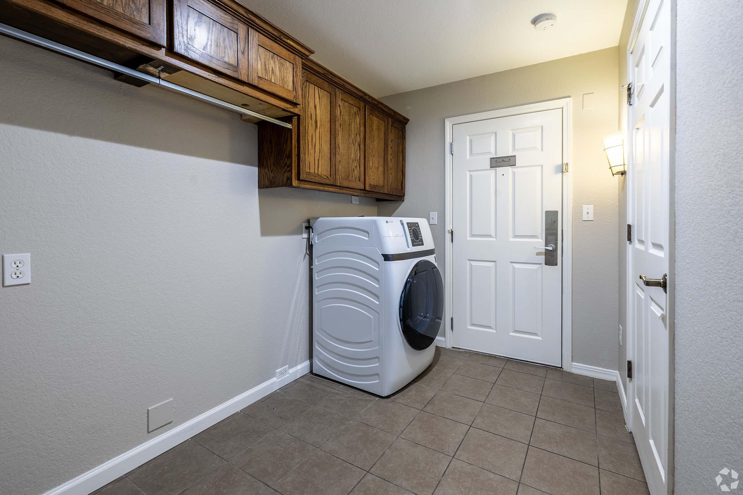 A clean laundry room featuring a modern white washing machine, wooden cabinets above for storage, and a door leading outside. There is a light fixture on the wall, and the floor is tiled, contributing to a functional and organized space.