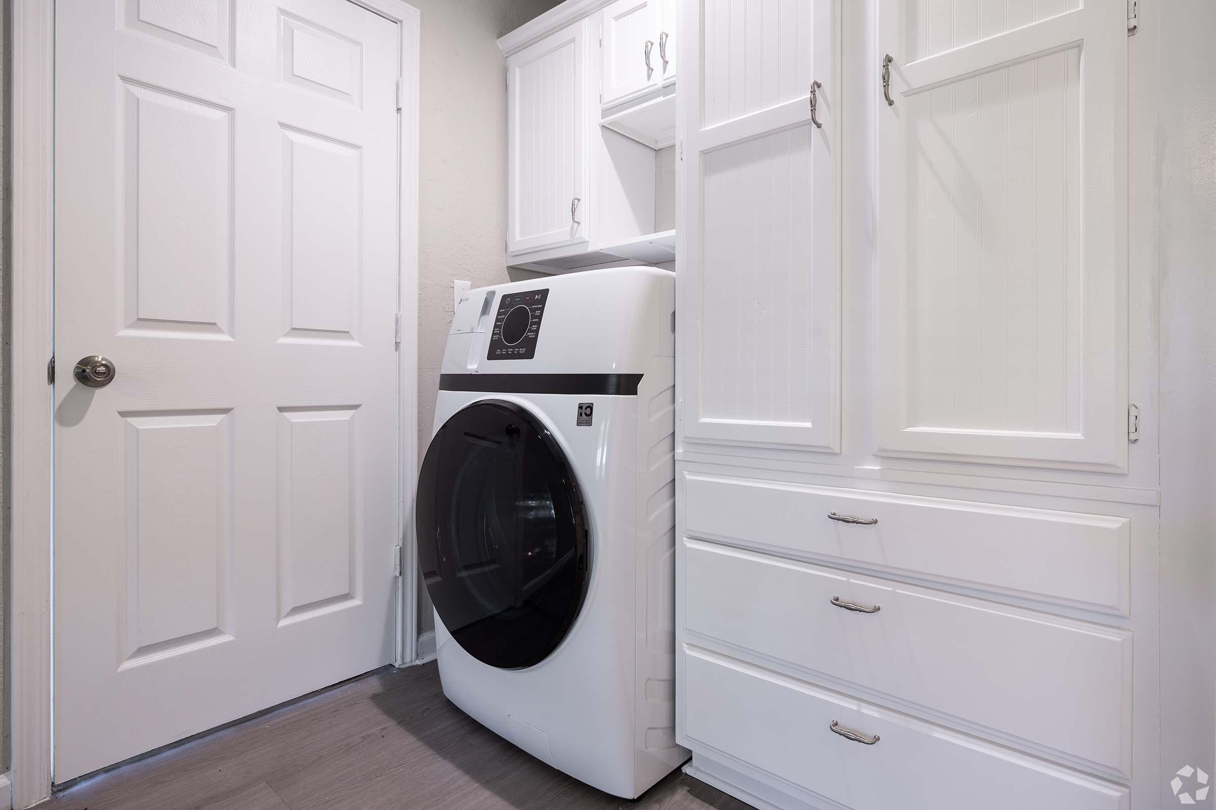 A clean laundry room featuring a modern front-loading washing machine in white, along with white cabinetry and drawers. A closed door is visible, and the room is well-lit, emphasizing a tidy and organized space.