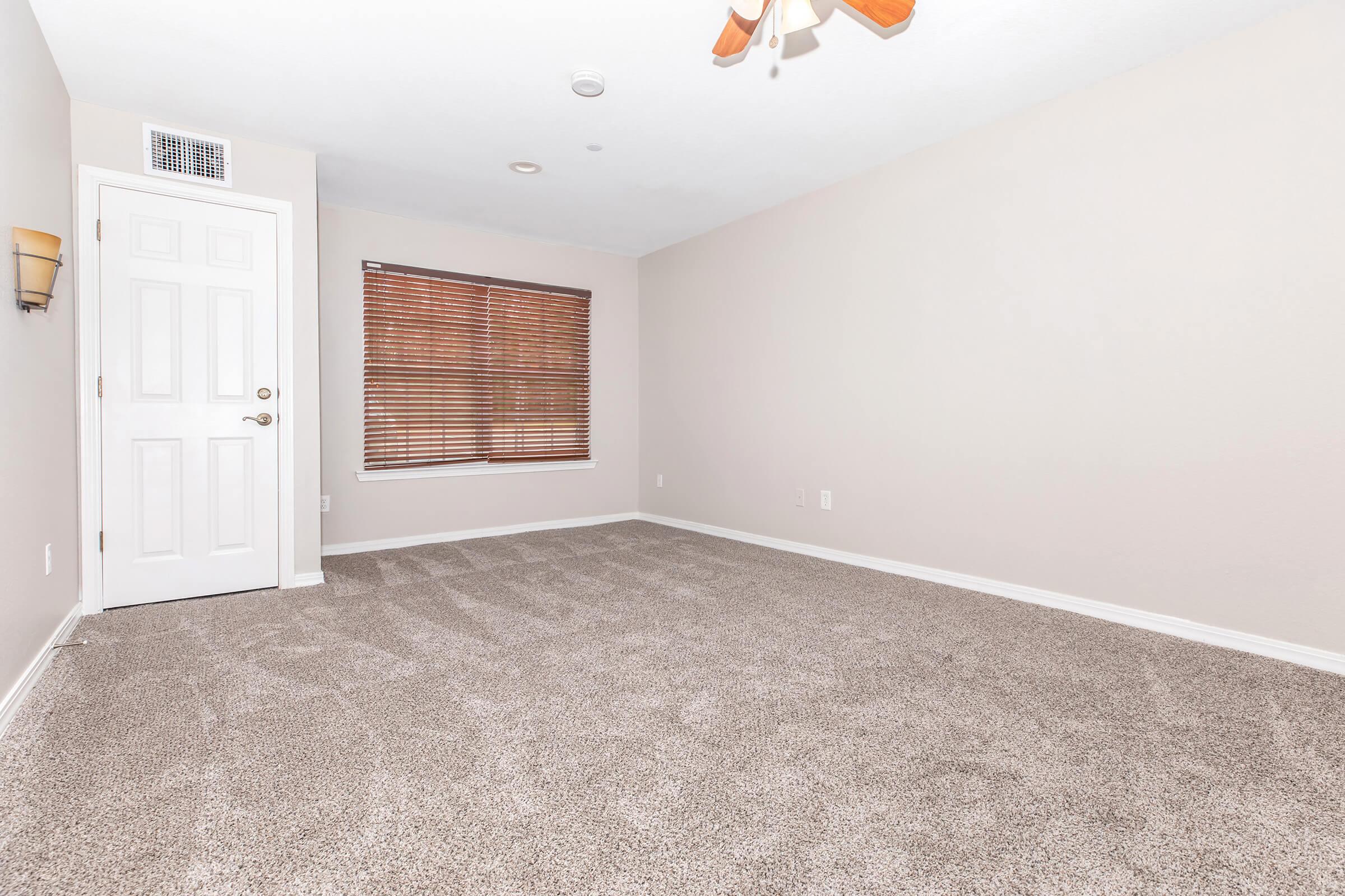 Empty room with light beige walls and carpeted flooring. A single window with blinds lets in natural light. The door is closed, and there is a wall sconce beside it. The ceiling features a ceiling fan, creating a spacious and minimalist atmosphere.