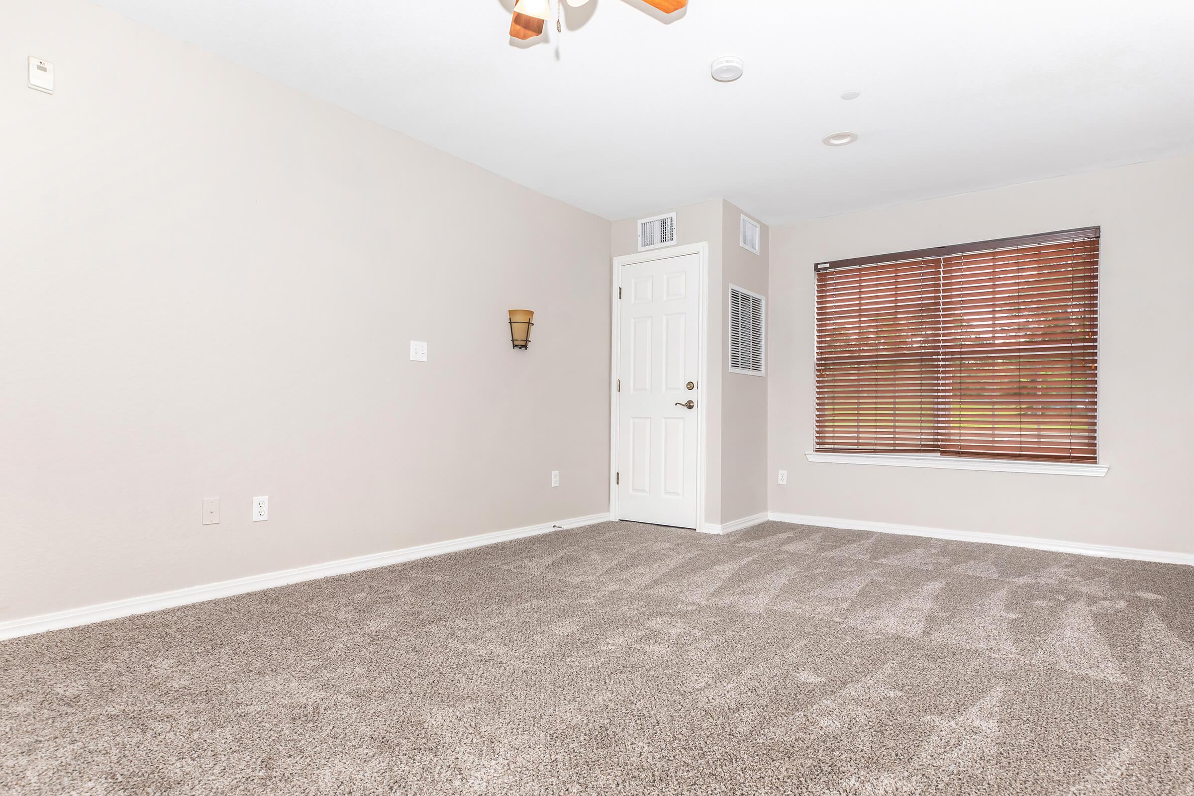 A spacious interior room with light beige walls and a plush gray carpet. It features a ceiling fan, a door with a window next to it, and a window with blinds allowing natural light. The room appears empty, creating an inviting, neutral space for potential furniture arrangement.