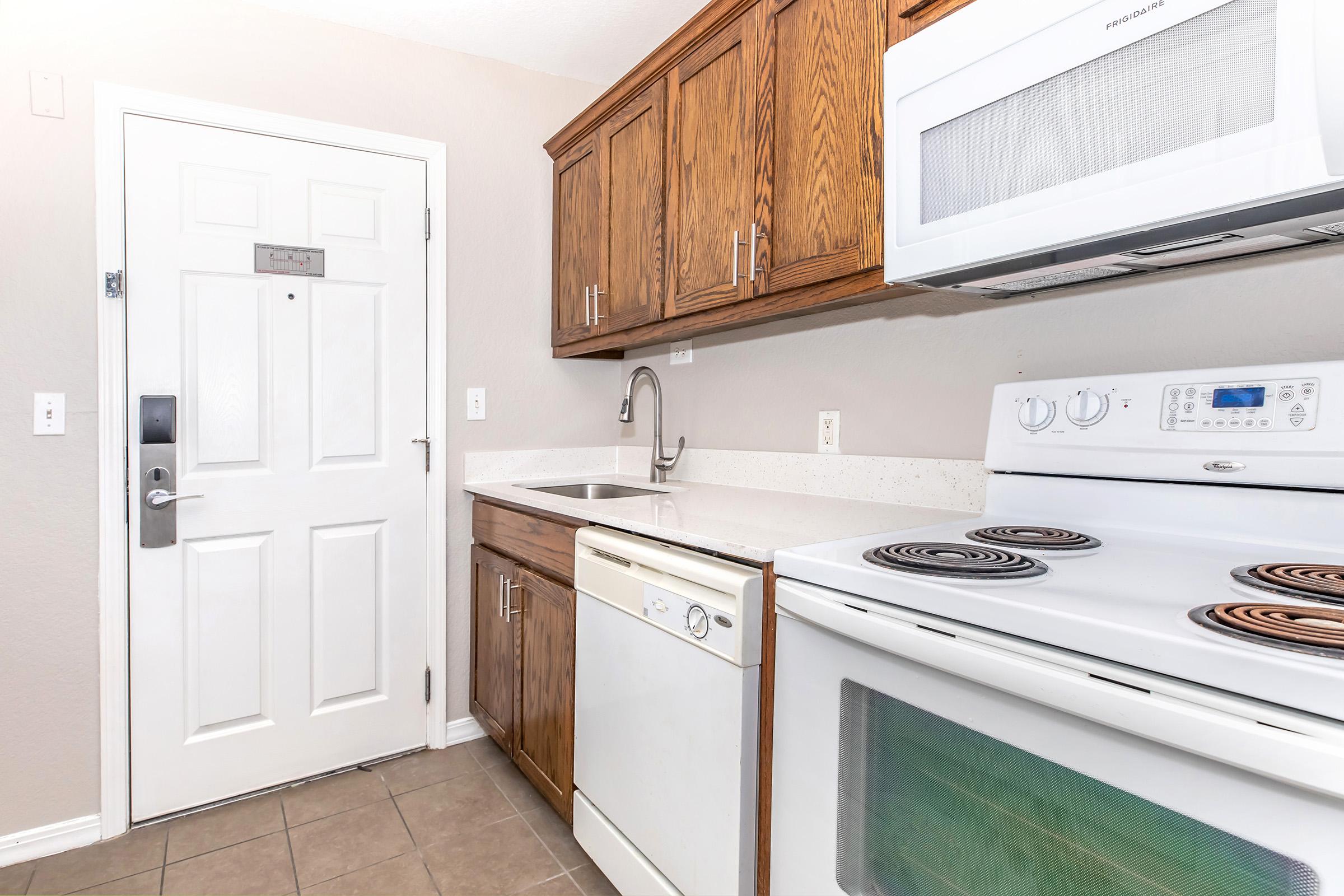 A small kitchen featuring wooden cabinets, a sink, a dishwasher, an electric stove, and a microwave. The entrance door is visible on the left with a key card locking system. The floor is tiled, and the overall color scheme is light and neutral.