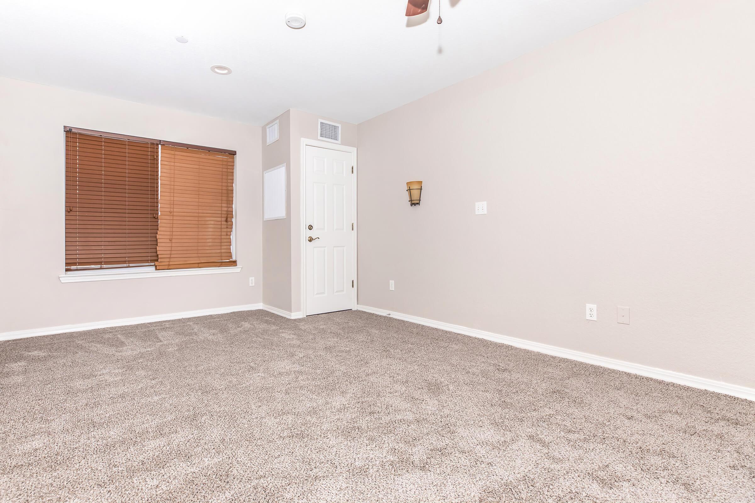 A spacious, empty room with beige walls and light carpeting. It features a window with wooden blinds, a wall sconce, and a door. The decor is minimal, creating a bright and open atmosphere, suitable for various uses.