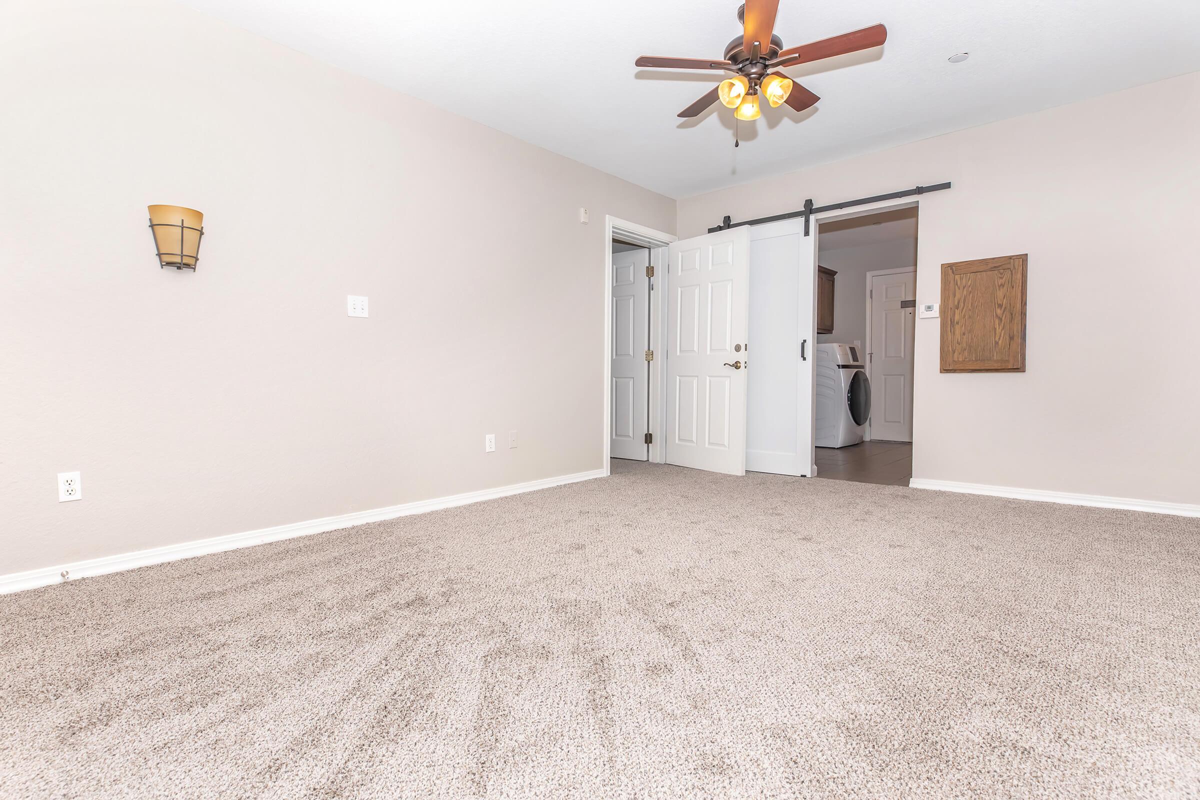 A spacious, neutral-colored room with beige carpet. There is a ceiling fan with wooden blades, a wall sconce on the left, and two closed doors leading to another room or hallway. A wooden panel is mounted on the right wall. The overall atmosphere is inviting and well-lit.