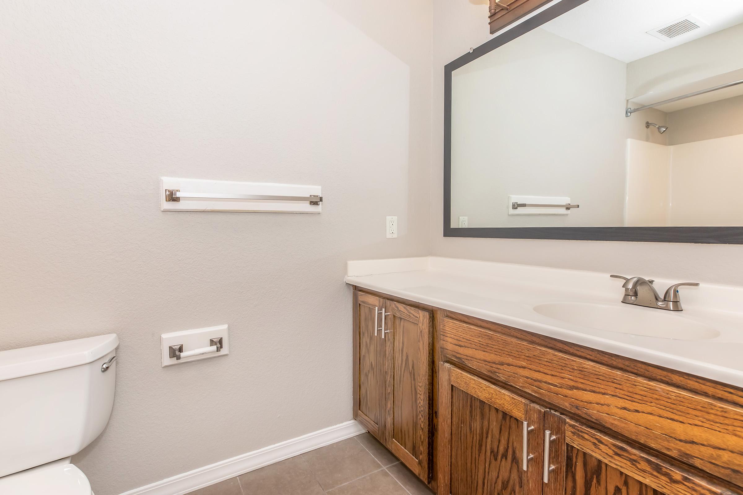 A clean bathroom featuring a white toilet, a wooden vanity with a sink, and a large mirror. The walls are painted a neutral color, and there is a towel bar mounted on the wall. The flooring is tiled in a light shade, contributing to a spacious and well-lit appearance.