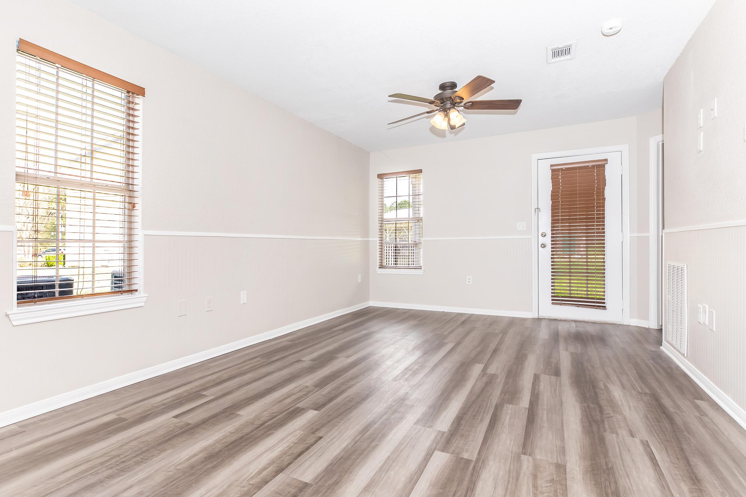 A spacious, empty room with light-colored walls and brown laminate flooring. It features two windows with blinds, a ceiling fan, and a door leading outside. The room is well-lit, creating a bright and inviting atmosphere.