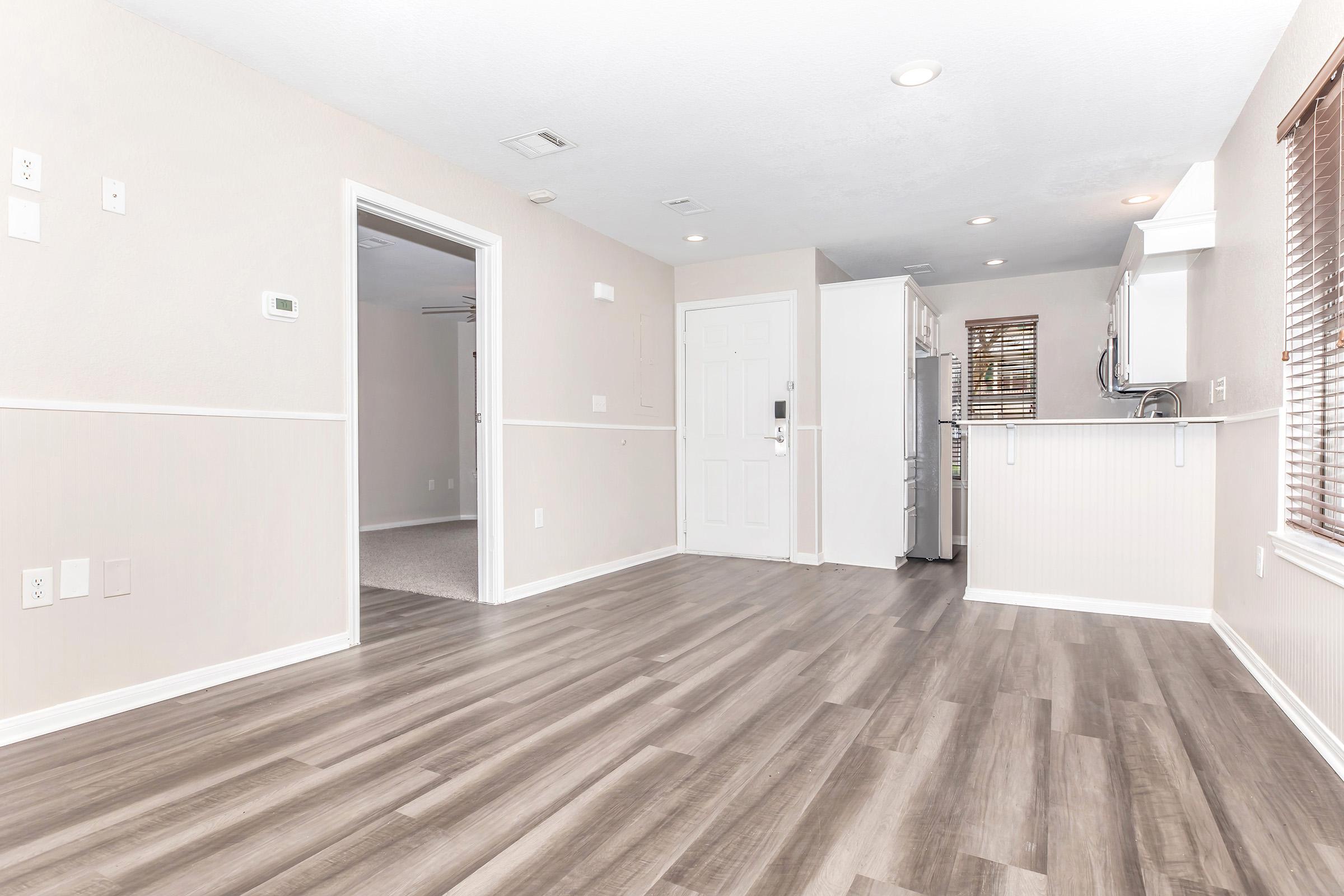 Bright, modern interior of a living space featuring light-colored walls, wood laminate flooring, and recessed lighting. An open doorway leads to another room, while the kitchen area is visible with white cabinetry and appliances. Large windows allow natural light to fill the room.