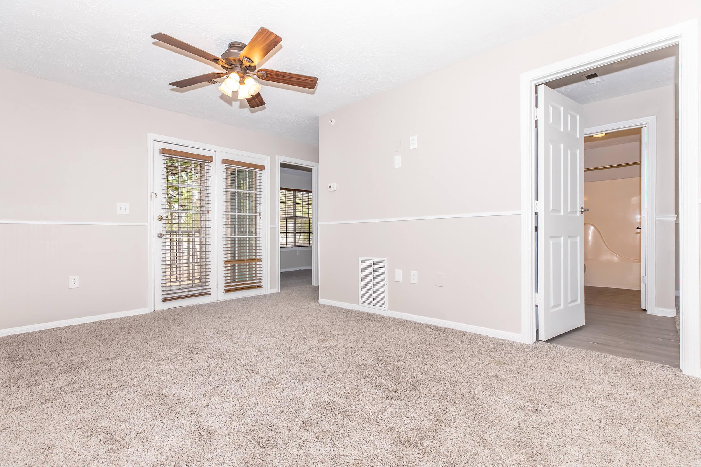 A bright, spacious living room with light-colored walls and soft carpet. A ceiling fan is visible, and there are double glass doors leading to an outdoor space. A doorway on the right leads to another room, while a wall vent adds to the home’s comfort. Natural light streams in, creating a welcoming atmosphere.