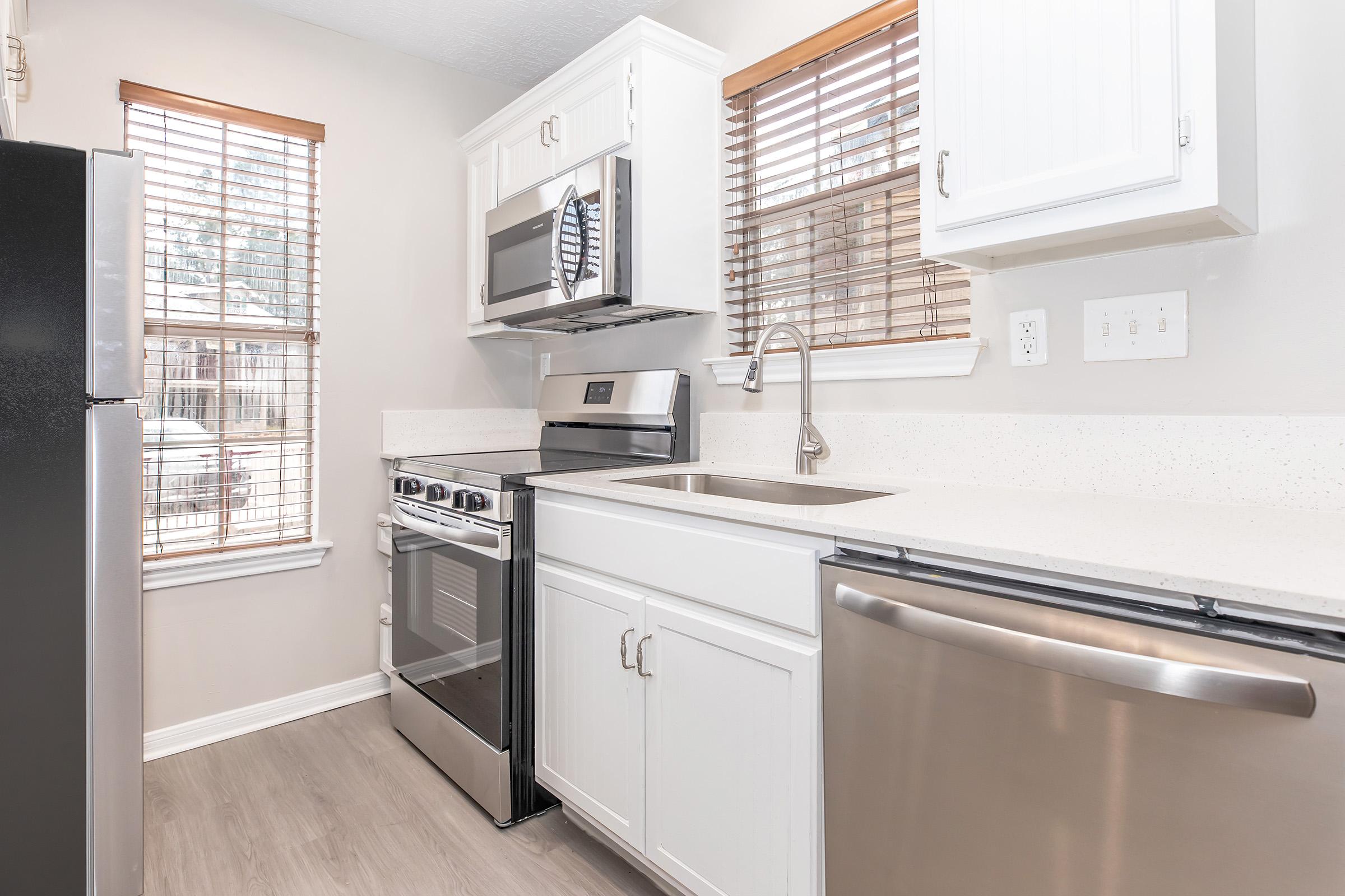Compact kitchen featuring white cabinets, stainless steel appliances, and a countertop. A black refrigerator, silver oven, and dishwasher are visible. Natural light comes through two windows with blinds, highlighting the modern design and clean aesthetic.