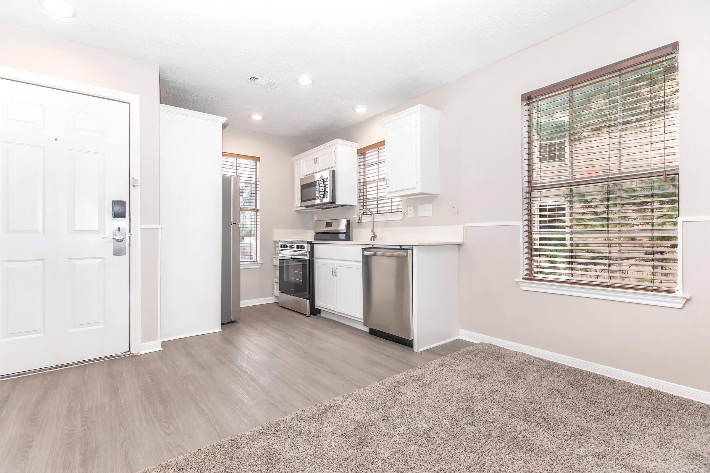 A modern kitchen featuring a stainless steel refrigerator, oven, and microwave, with white cabinets and a countertop. Natural light filters through window blinds, illuminating the space. The flooring is light wood, and there's a plush carpet in front of the entrance door.