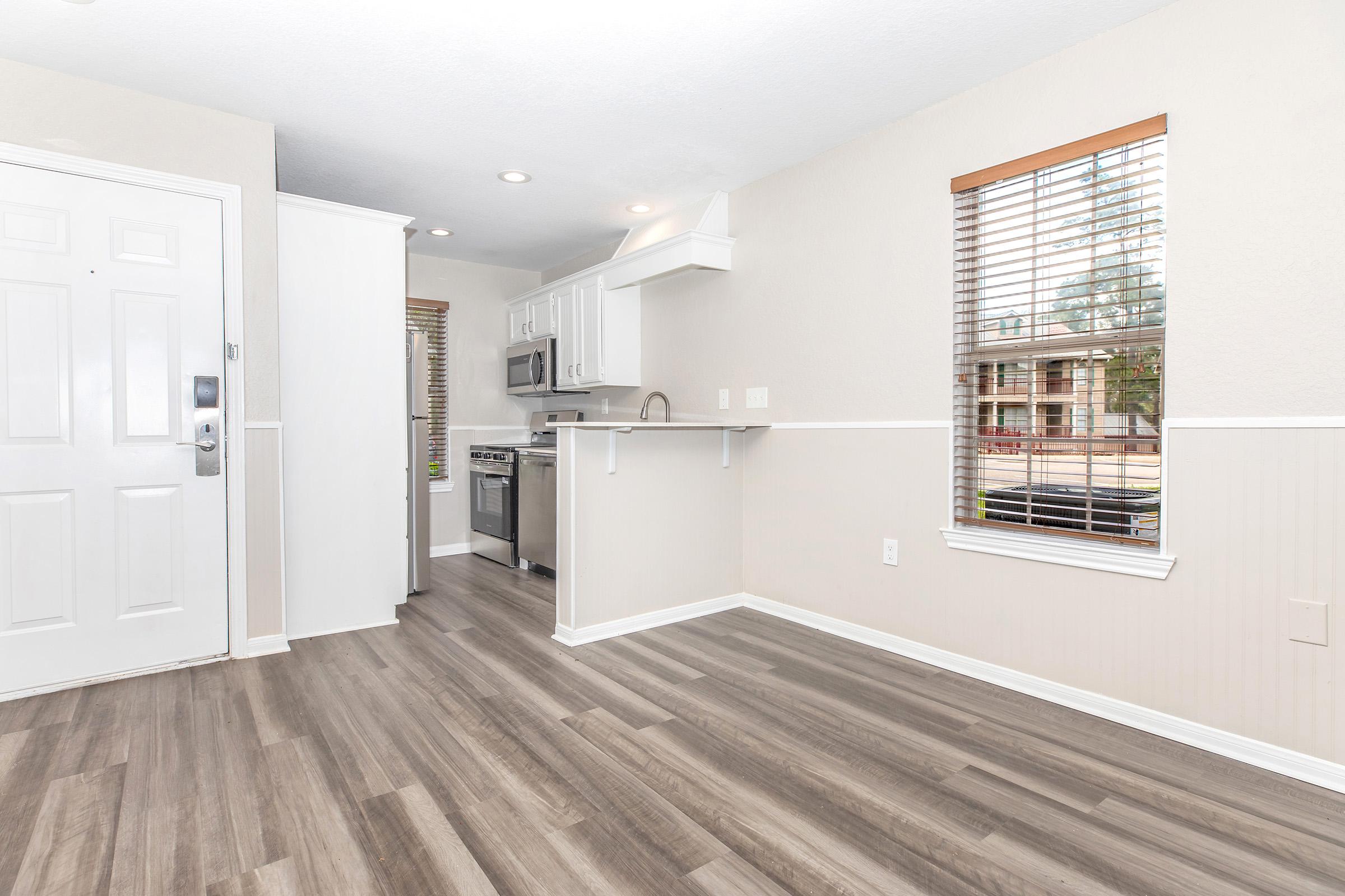 A well-lit, modern living space featuring light-colored walls and sleek laminate flooring. To the left, there's a white front door with a small window beside it. The kitchen area is visible in the background with stainless steel appliances, and a window with wooden blinds lets in natural light.