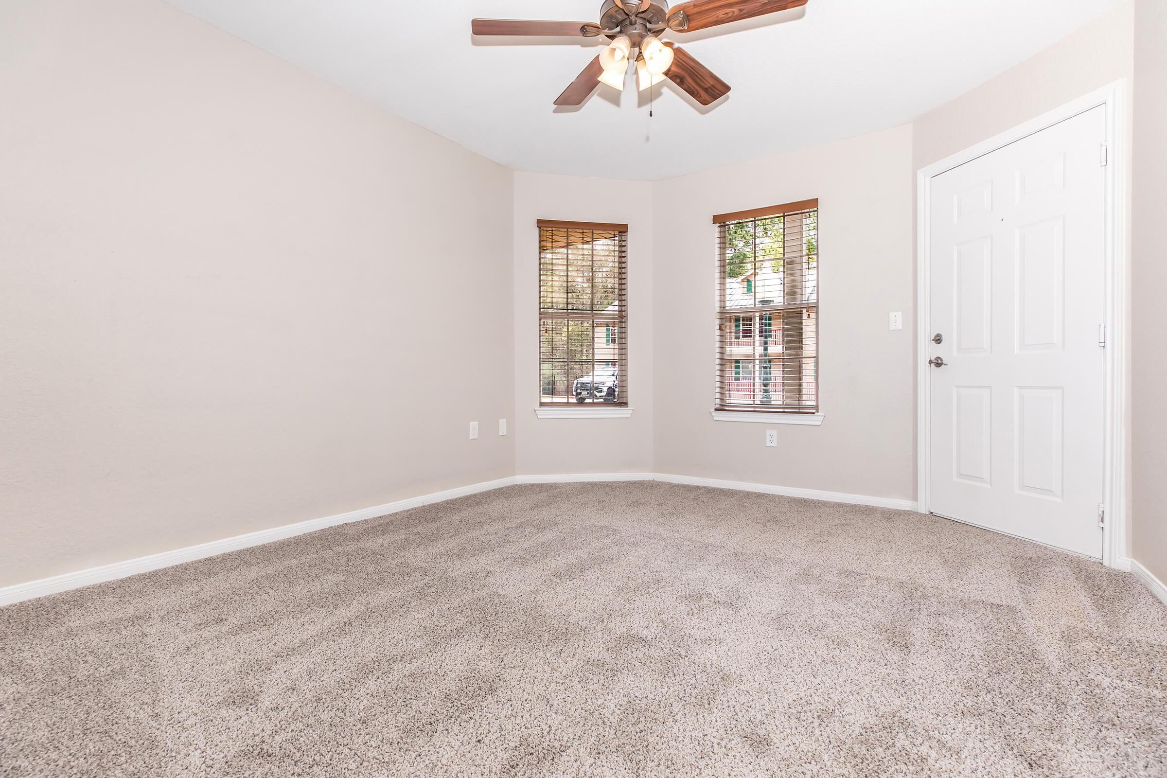 A vacant room with light beige walls and soft, beige carpet. It features two windows with blinds and a ceiling fan with wooden blades. The entry door is on the right, and the room has a cozy, empty feel, suitable for various uses. Natural light brightens the space.