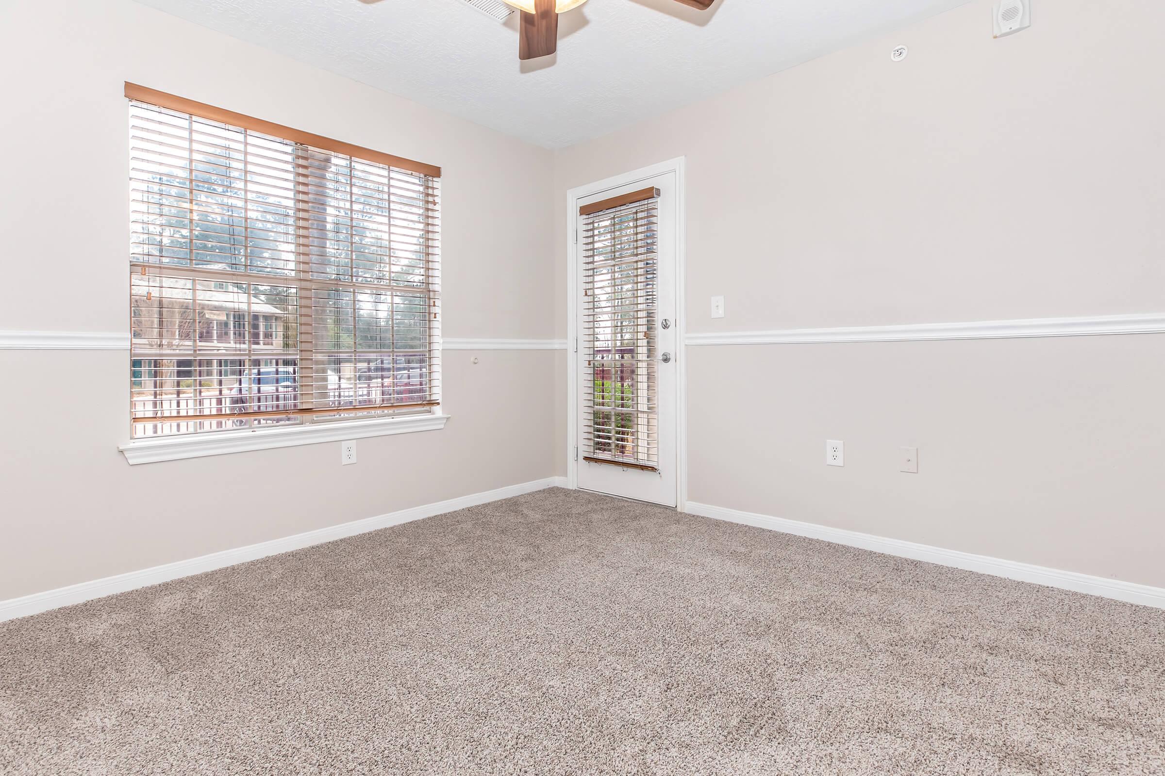 A well-lit empty room with beige walls and carpet, featuring a ceiling fan. There is a window with wooden blinds providing natural light, and a door with vertical blinds leads outside. The space has a clean and inviting appearance, suitable for various uses.