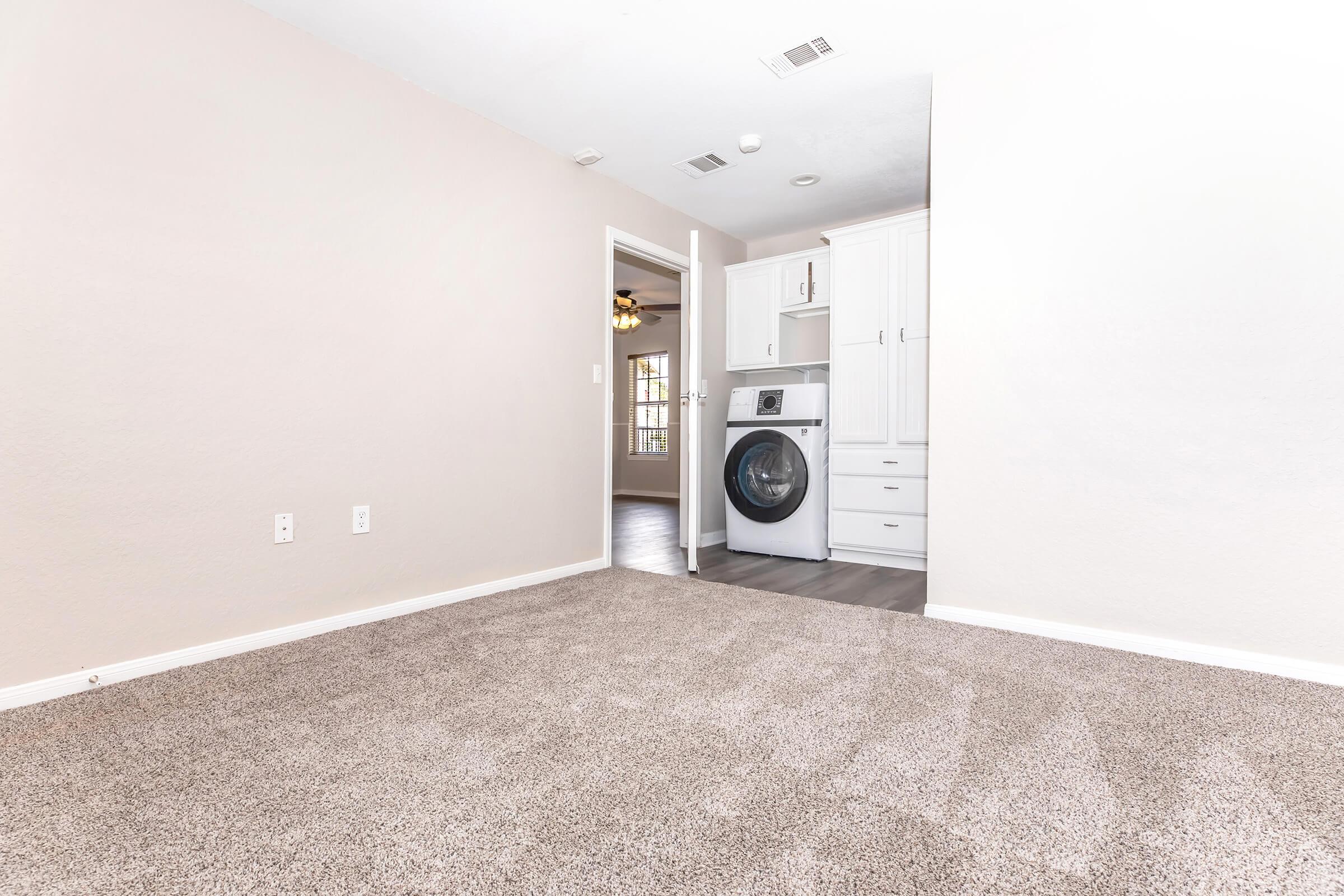 A spacious room featuring light-colored walls and plush carpeting. On one side, there's a laundry area with a washer and dryer, and storage cabinets. A doorway leads to another room with a window and ceiling fan, enhancing the overall modern and airy feel of the space.