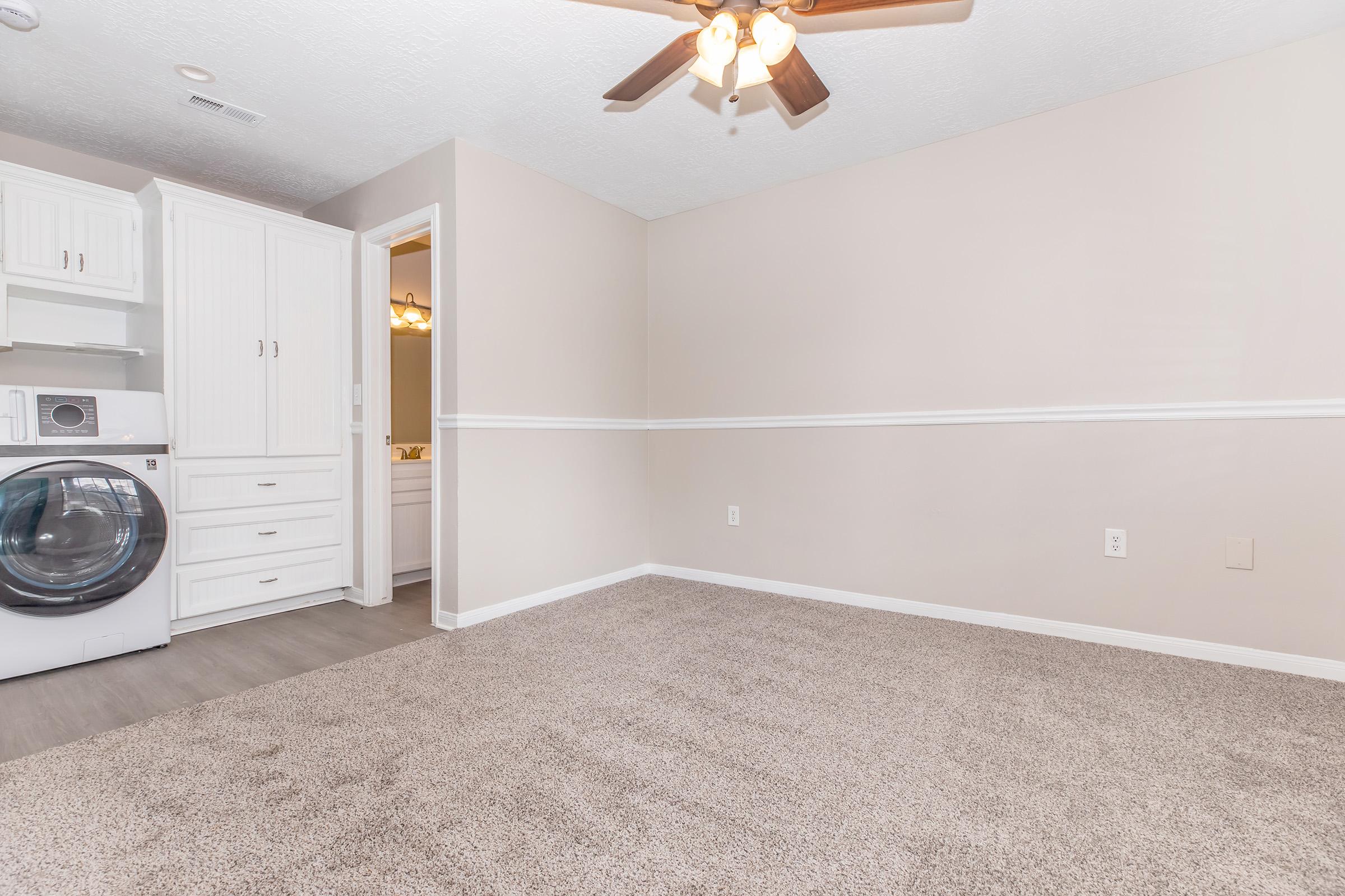 A cozy room with light beige walls and carpet, featuring a ceiling fan. On the left, there is a laundry machine, and a doorway leads to a bathroom. The space is clean, with a simple and neutral aesthetic.