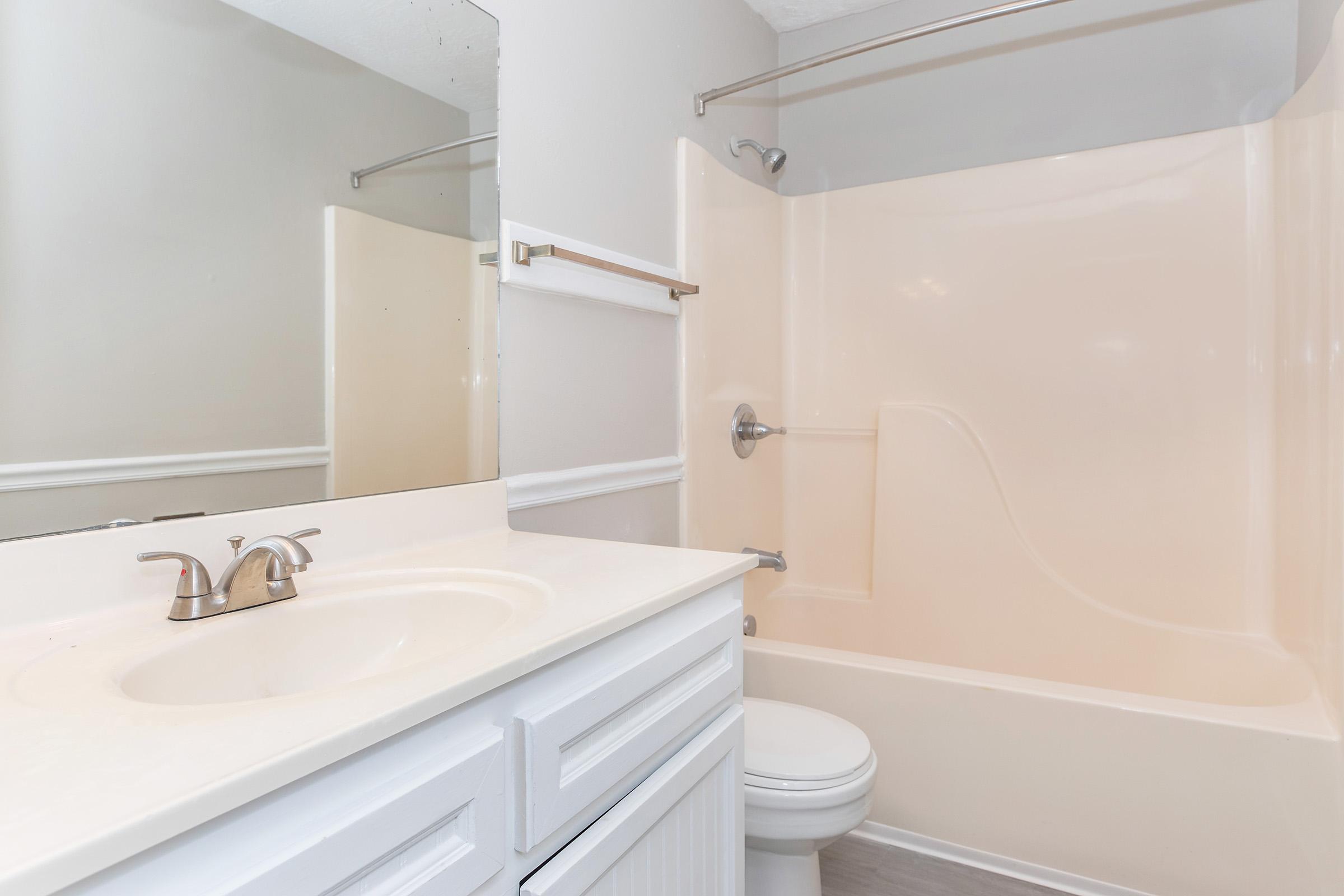 A simple bathroom featuring a shower-tub combo, a white vanity with a sink, and a mirror. The walls are painted light gray, and the flooring is a light neutral color. The overall design is clean and minimalistic.