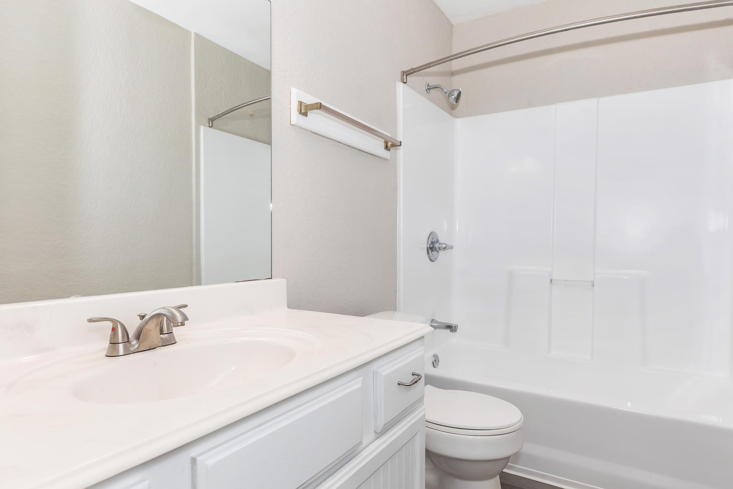 Clean, modern bathroom featuring a white bathtub and shower combination, a sink with a single faucet, a large mirror, and light-colored walls. The space includes a towel bar next to the mirror and a toilet positioned beside the shower. The overall design is bright and minimalistic.