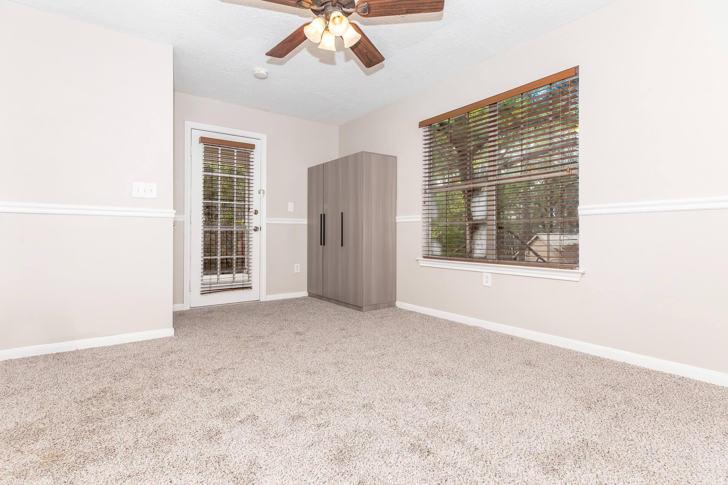 A clean, empty room with light beige walls and a textured carpet. Natural light filters through a window with horizontal blinds, and there's a door leading outside. A modern gray cabinet stands against one wall, creating a minimalist and neutral aesthetic.