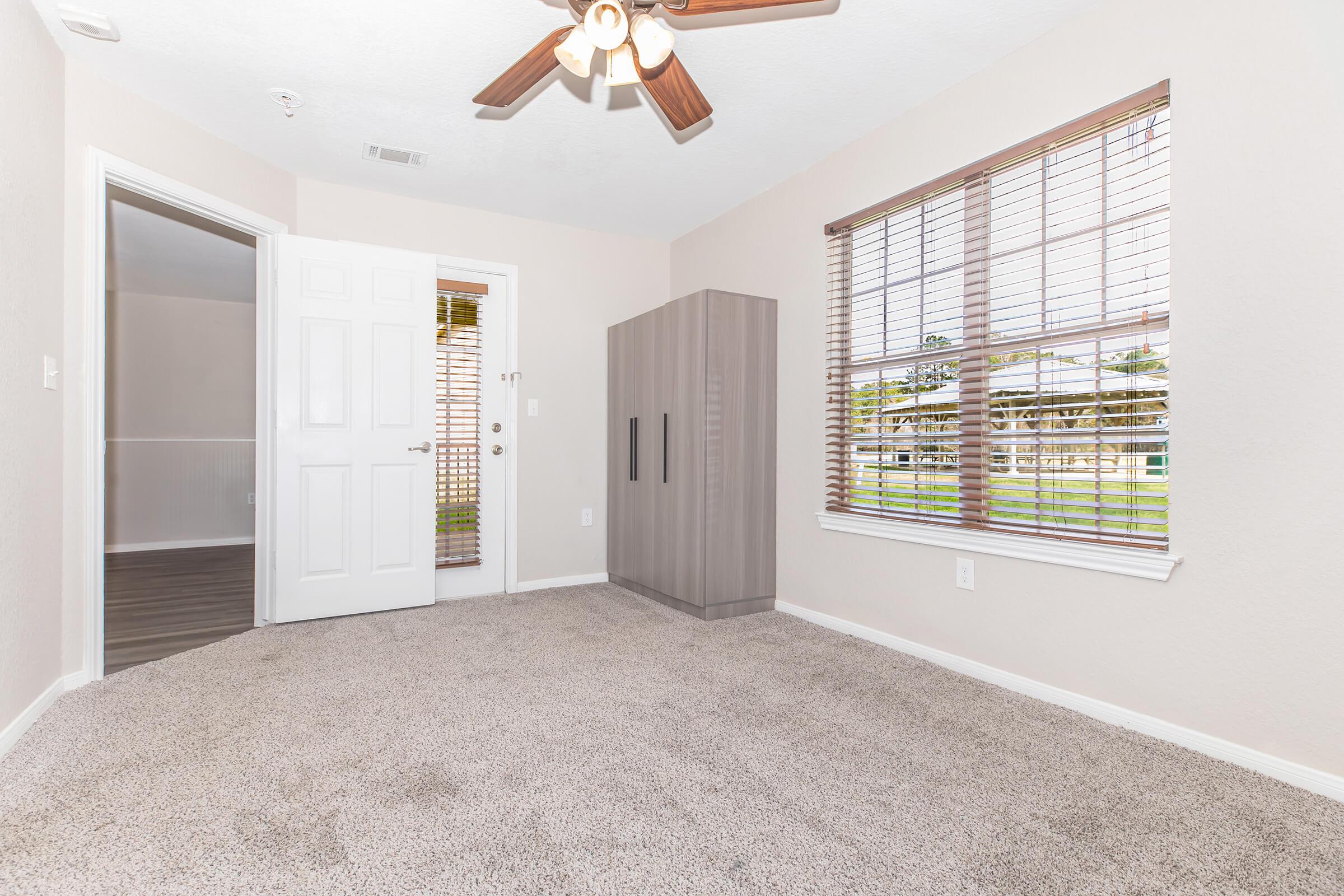 A well-lit room featuring a ceiling fan, light beige walls, and carpeted flooring. There is a door leading outside, a closet with sliding doors, and a window with wooden blinds providing a view of greenery outside. The room has a modern, minimalist aesthetic.