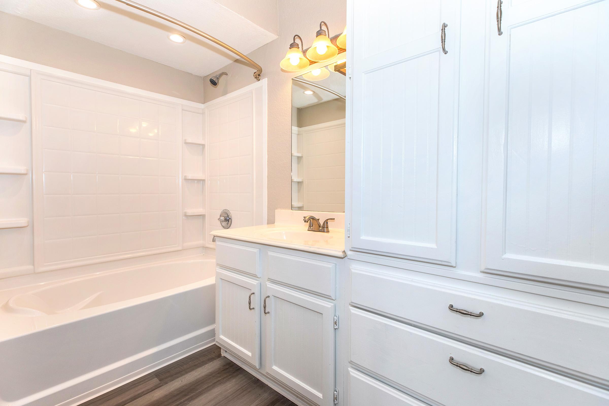 A bright and modern bathroom featuring a white bathtub with a shower, white cabinetry with silver handles, and a large mirror above the sink. The walls are painted in a neutral tone, and there are overhead lights illuminating the space, creating a clean and inviting atmosphere.