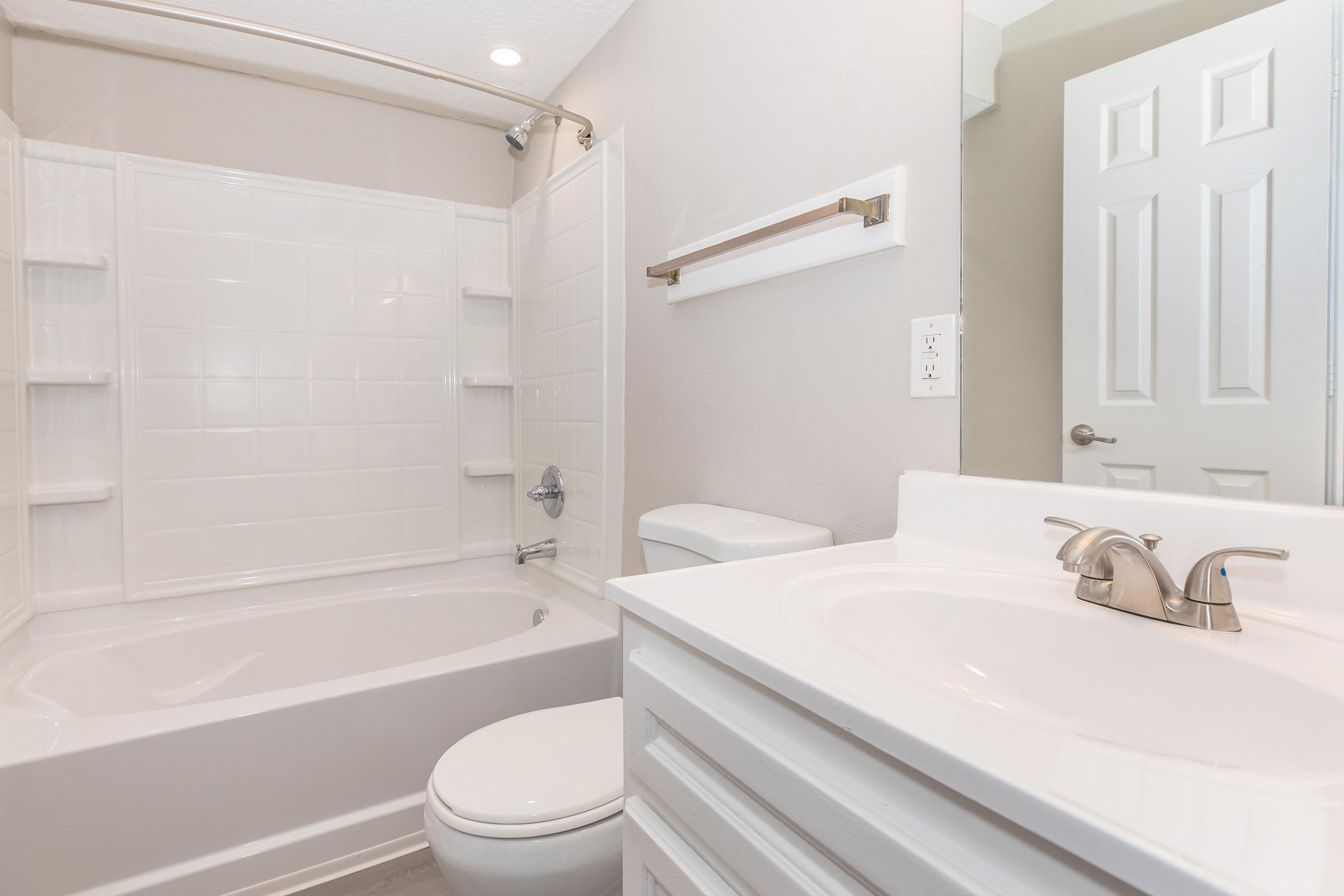 A clean and modern bathroom featuring a white bathtub with a shower, a toilet, and a sink with a faucet. The walls are painted in a light color, and there are shelves in the shower area for toiletries. A door leads to another room, and the overall decor is simple and contemporary.