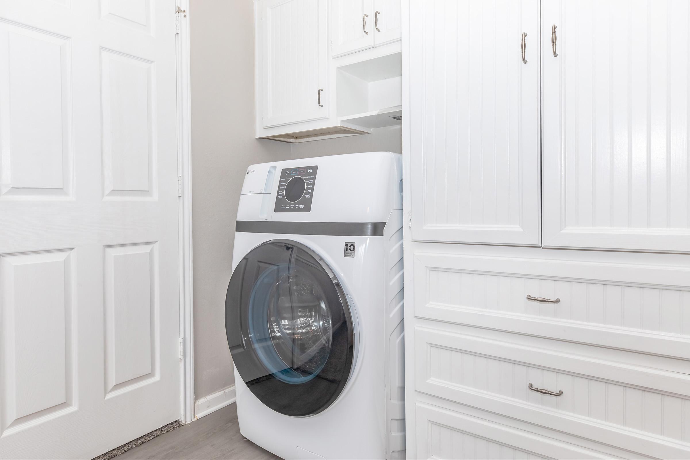 A modern front-loading washing machine in a laundry room, positioned between white cabinetry and a door. The cabinetry features a sleek design with a light finish, while the washing machine has a digital control panel and a transparent door, showcasing its interior.