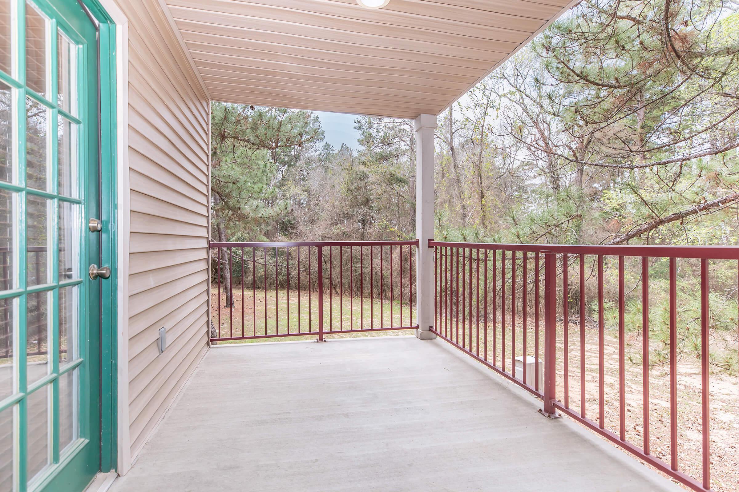 A spacious balcony with a metal railing, overlooking a wooded area. The porch features light gray flooring and tan siding on the house. A green door is visible, leading into the interior. The atmosphere is calm and serene, surrounded by greenery and trees.