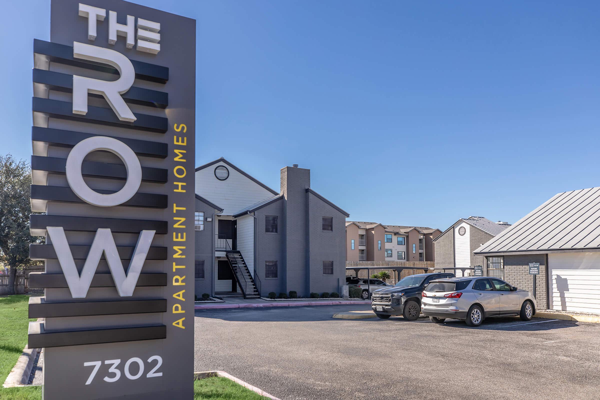 Sign for The Row Apartment Homes featuring large, stylized letters. In the background, there are two apartment buildings with a clear blue sky. Cars are parked in front of the buildings, and the area is surrounded by green grass. The sign includes the address 7302 prominently displayed.