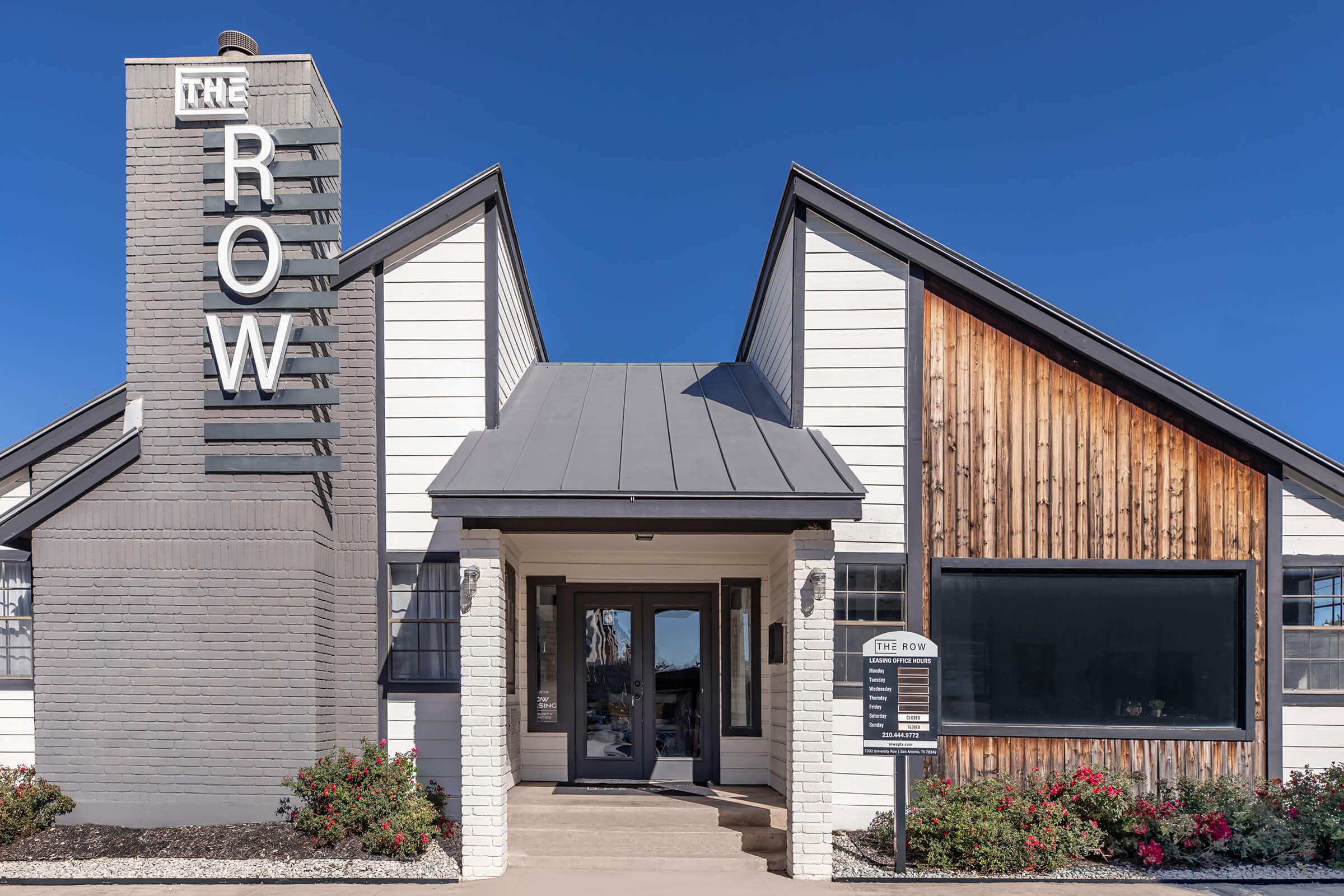 Exterior of a modern building featuring a combination of white and wooden siding. A large sign reading "THE ROW" is prominently displayed on the left. The entrance is framed by large windows, with landscaped flower beds on either side. Clear blue sky in the background.
