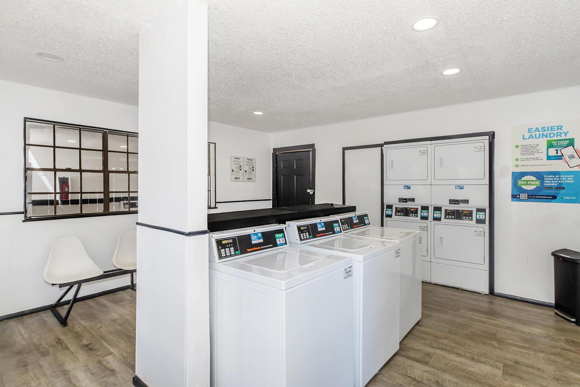 A clean and modern laundry room featuring multiple washing machines and dryers, a waiting area with sleek white chairs, and informational signage on the walls. The space is well-lit with a neutral color scheme and a polished floor.
