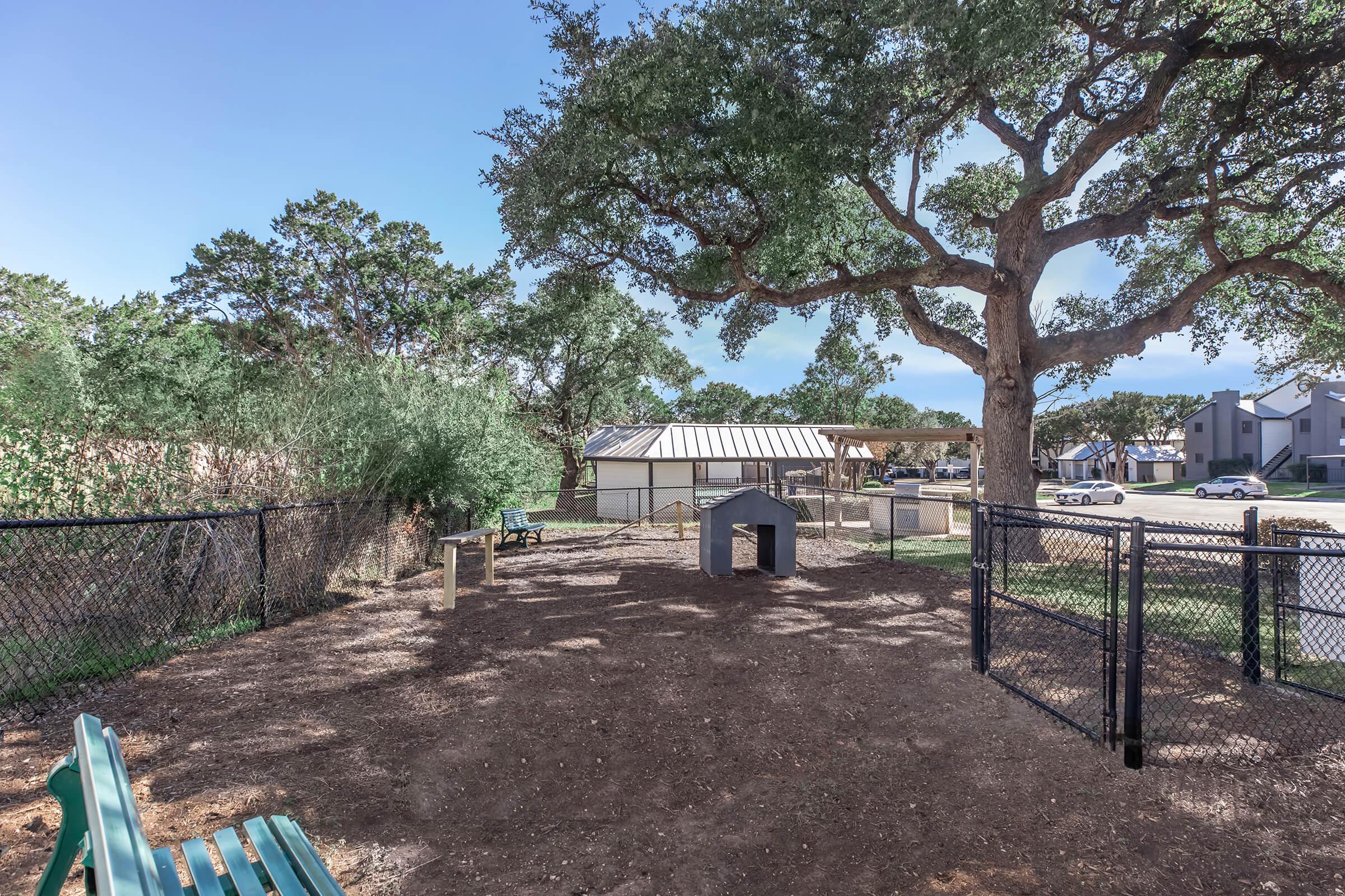 A spacious dog park with a fenced area featuring a small doghouse, shaded by a large tree. Nearby are benches for seating and a view of a well-maintained landscape with a building in the background. The ground is mostly dirt, suitable for play.