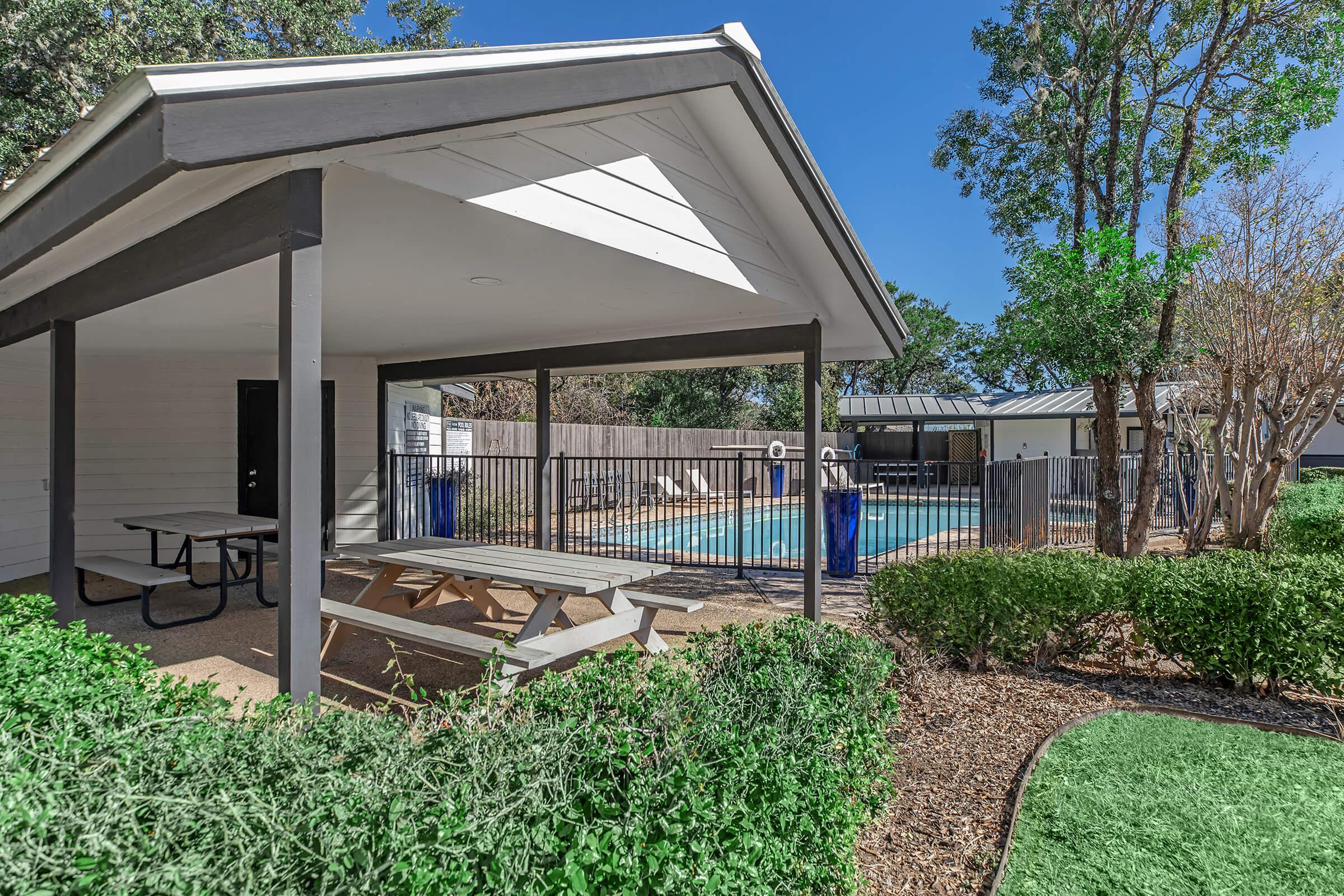 A shaded gazebo with picnic tables overlooks a fenced swimming pool surrounded by trees and shrubs. The pool area features a diving board and a clear blue sky above, creating a relaxing outdoor space for gatherings and leisure activities.