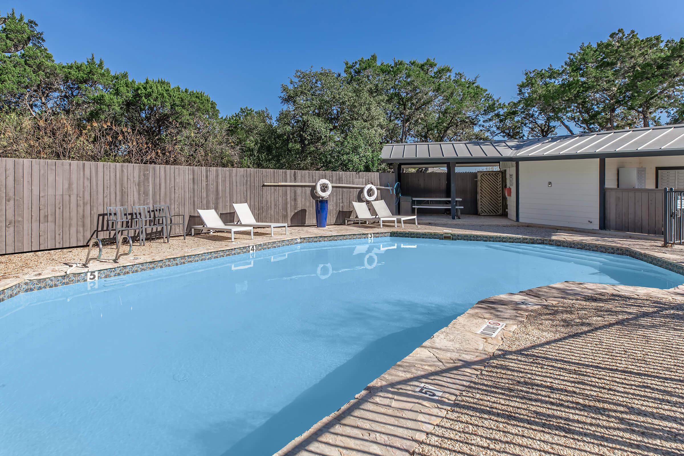 A serene outdoor pool area featuring a clear blue water pool surrounded by lounge chairs. There’s a wooden fence in the background, a small shed with a covered area, and trees providing shade nearby. The sky is bright and clear, creating a relaxing atmosphere.