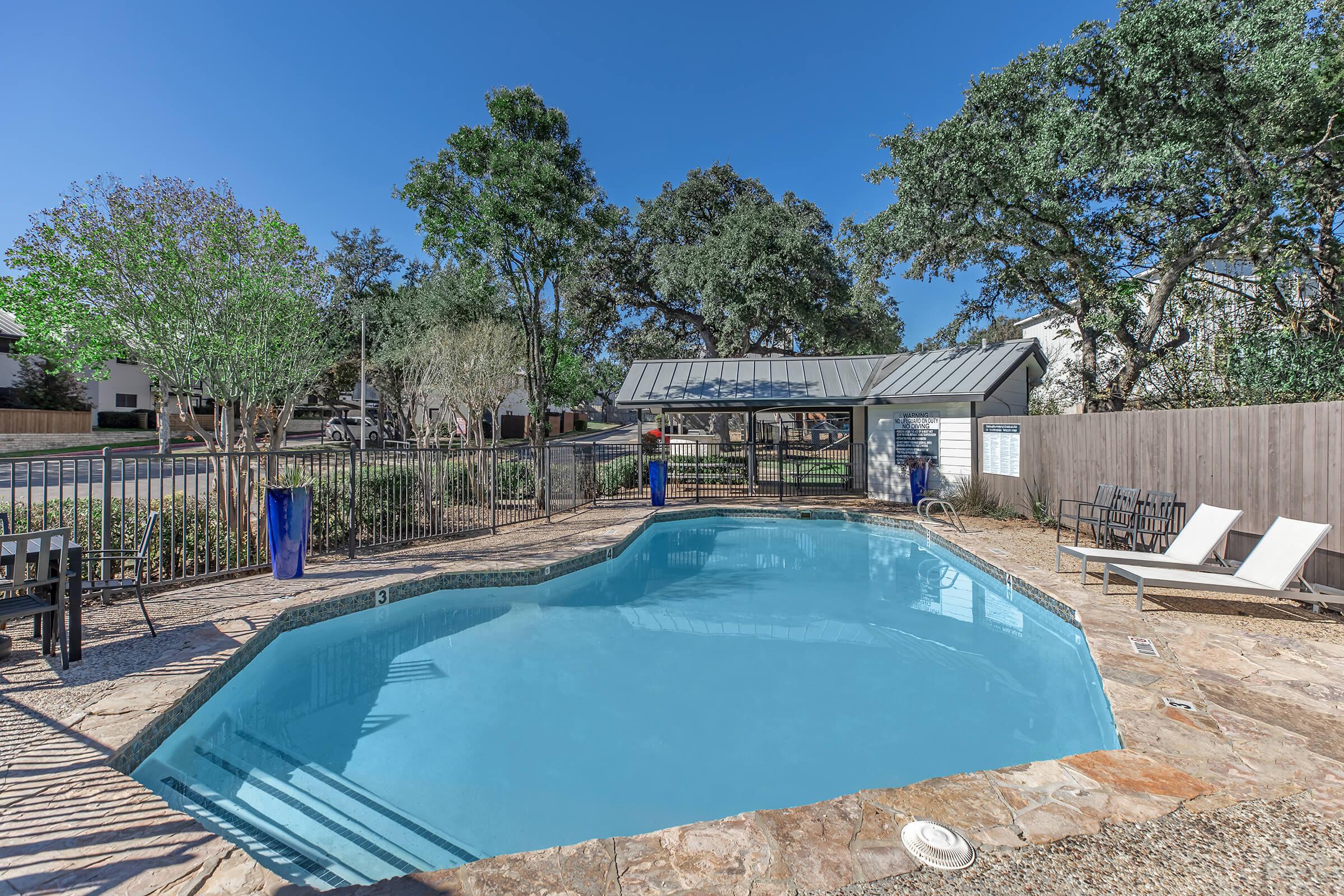 A clear blue swimming pool surrounded by a stone patio, with lounge chairs positioned nearby. Lush green trees and shrubs are visible in the background, along with a shaded outdoor seating area and a fenced perimeter. The sky is bright and clear, indicating a sunny day.