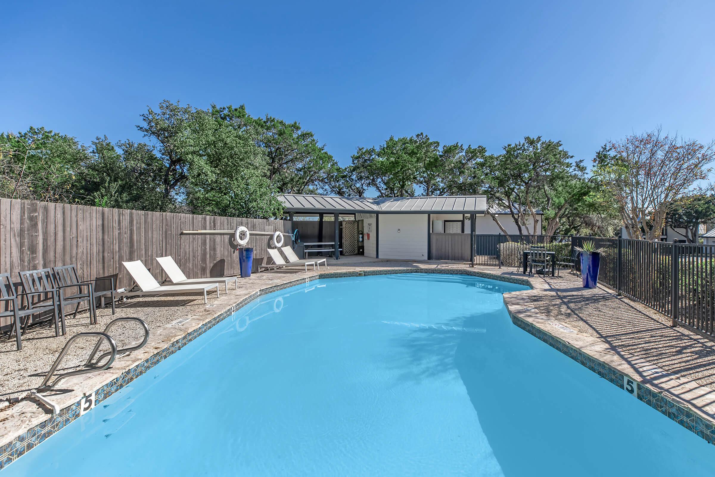 A clear blue swimming pool surrounded by lounge chairs and shaded by trees. A wooden fence provides privacy, and a building is visible in the background, featuring a porch area. The sky is bright blue, indicating a sunny day.
