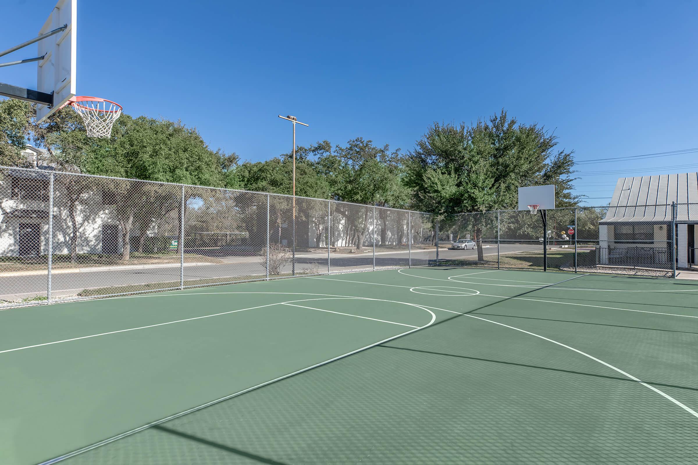 View of an outdoor basketball court enclosed by a chain-link fence. The court features a basketball hoop at each end, green asphalt surface, and surrounding trees under a clear blue sky. A nearby building is visible in the background.
