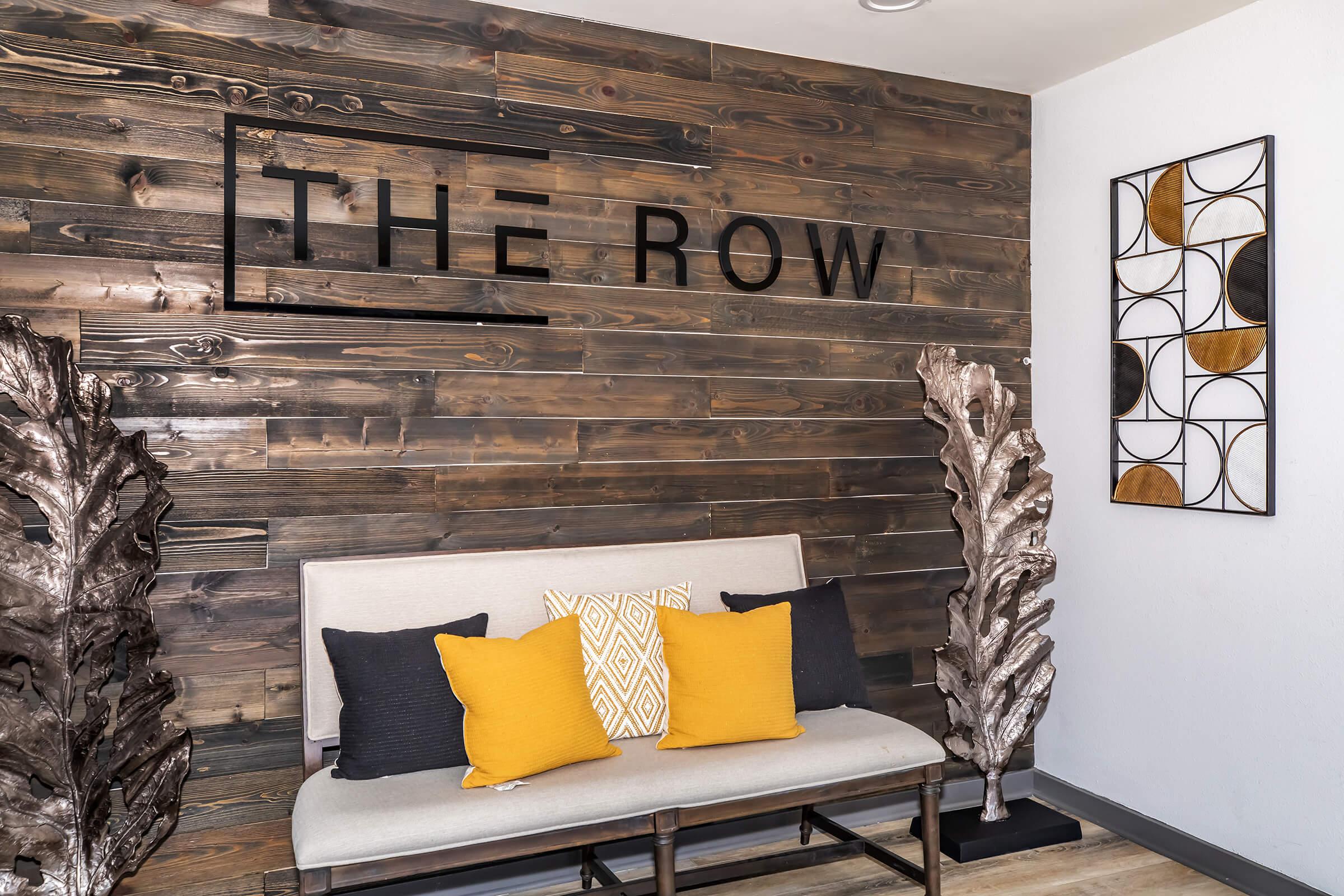 A cozy interior space featuring a wooden accent wall with the text "THE ROW" in bold letters. A stylish bench with light gray cushions is adorned with yellow and black pillows. To the right, an abstract wall art piece with geometric shapes and warm colors adds a decorative touch.