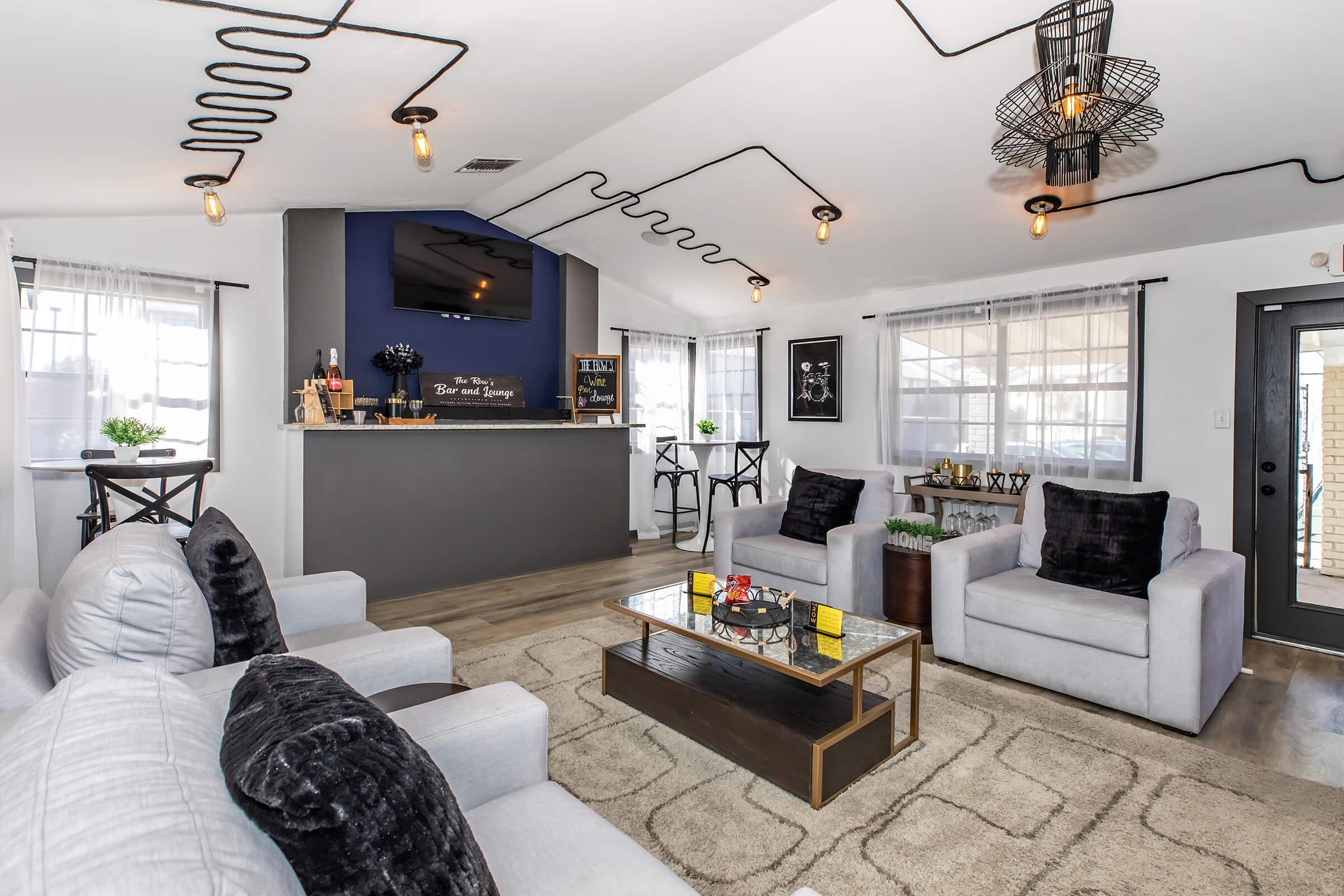 A modern, stylish living room featuring two gray sofas with black accent pillows, a glass coffee table, and a bar area in the background. The space has industrial lighting, light wood flooring, and large windows with sheer curtains, creating a bright and inviting atmosphere.