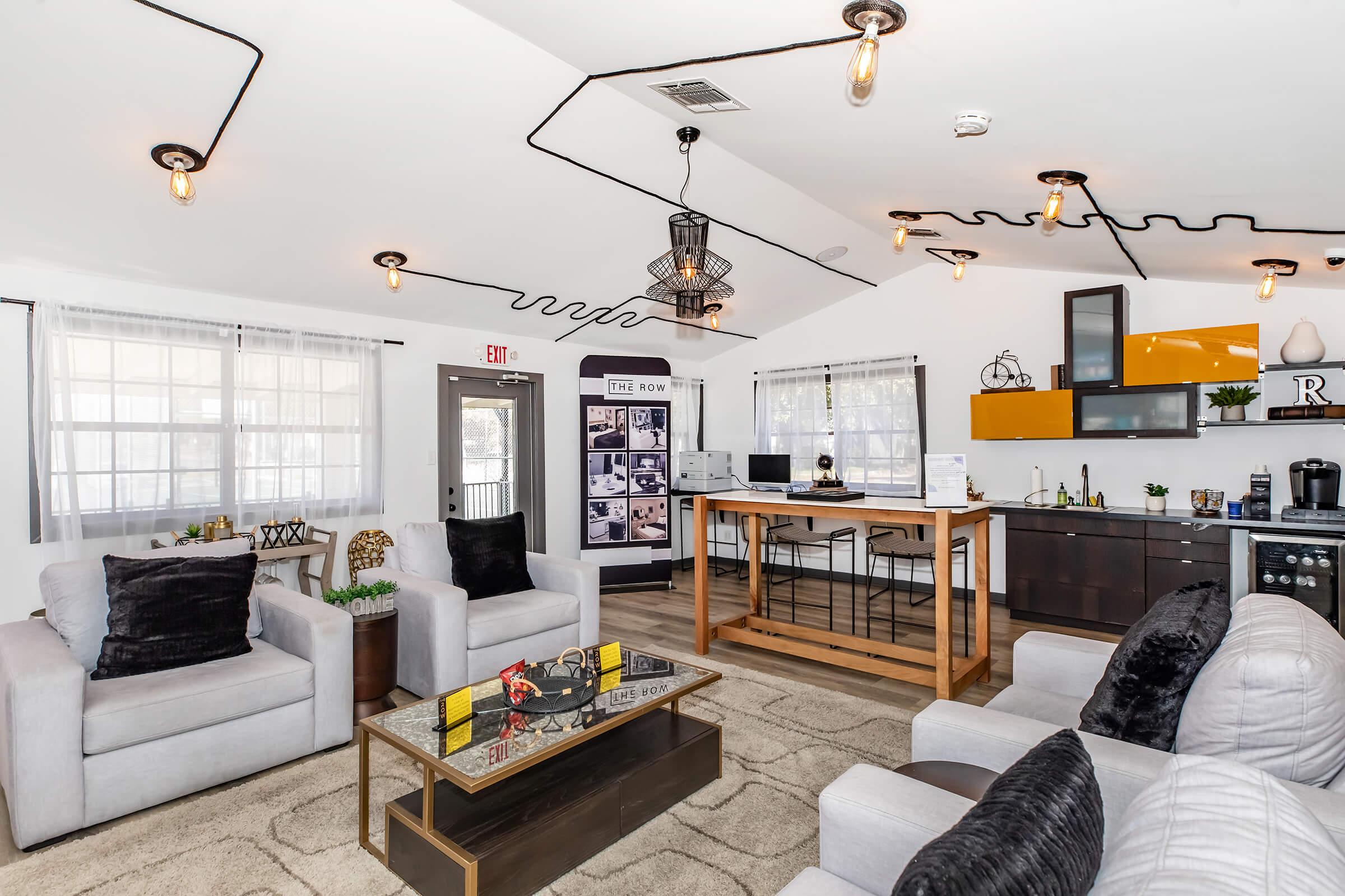 A modern living space featuring two white armchairs and a gray sofa arranged around a stylish coffee table. The room includes a kitchen area with sleek cabinetry, bright lighting fixtures, and decorative elements. Large windows allow natural light to fill the space, creating a warm, inviting atmosphere.