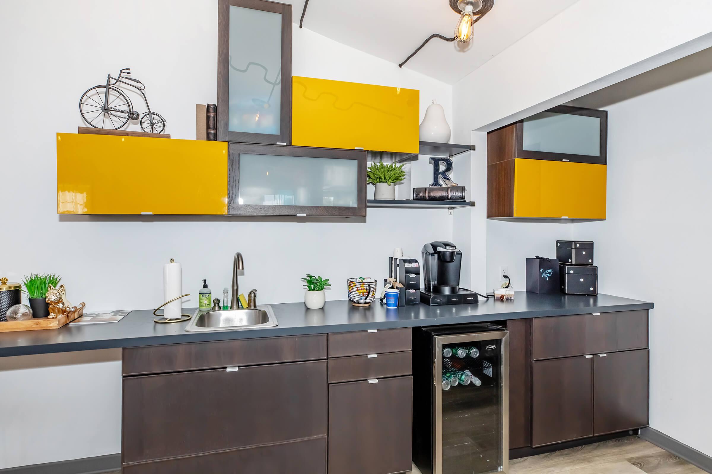 A modern kitchen with dark cabinetry and bright yellow upper cabinets, featuring a stainless steel sink, coffee machine, and mini fridge. Decor elements include plants and kitchen utensils, complemented by a stylish light fixture overhead. The space has a contemporary, inviting design.