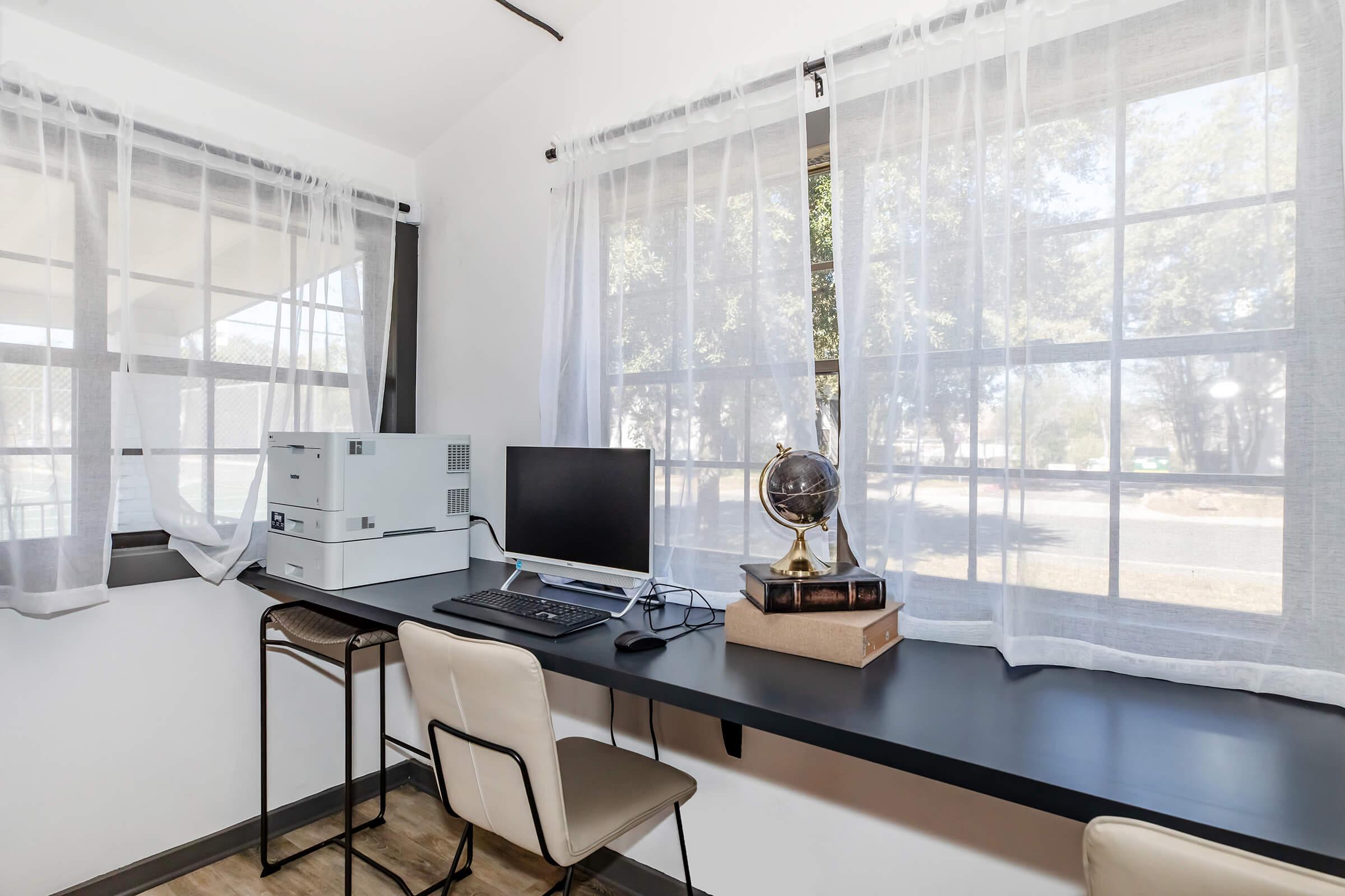 A bright home office space featuring a desk with a computer monitor, a printer, and a decorative globe. Large windows with sheer white curtains allow natural light to illuminate the room, while a minimalist chair completes the setup. The decor is simple yet inviting, ideal for productivity.