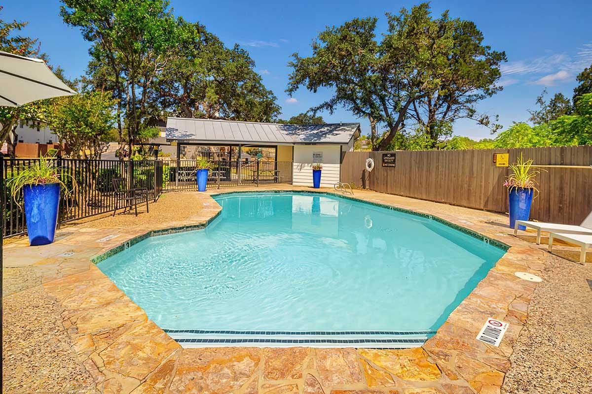 A clear swimming pool surrounded by lush greenery and trees. The pool features a stone deck and blue planters on either side. A shaded seating area is visible in the background, complete with a roofed pavilion and fencing for safety. The bright sky adds to the inviting atmosphere of the outdoor space.