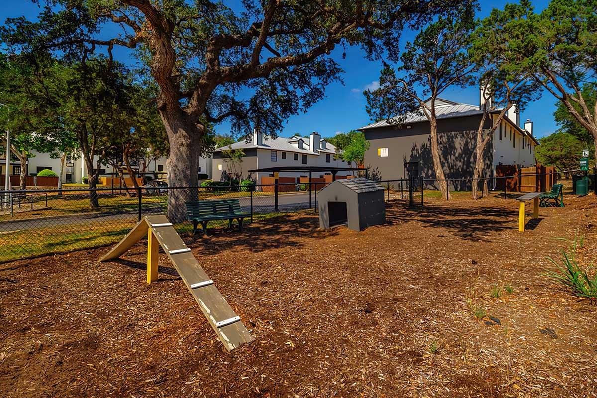 A sunny outdoor dog park features a wooden ramp, benches, and a doghouse, surrounded by trees and a fence. Nearby residential buildings are visible in the background. The ground is covered with mulch, creating a natural play area for pets.