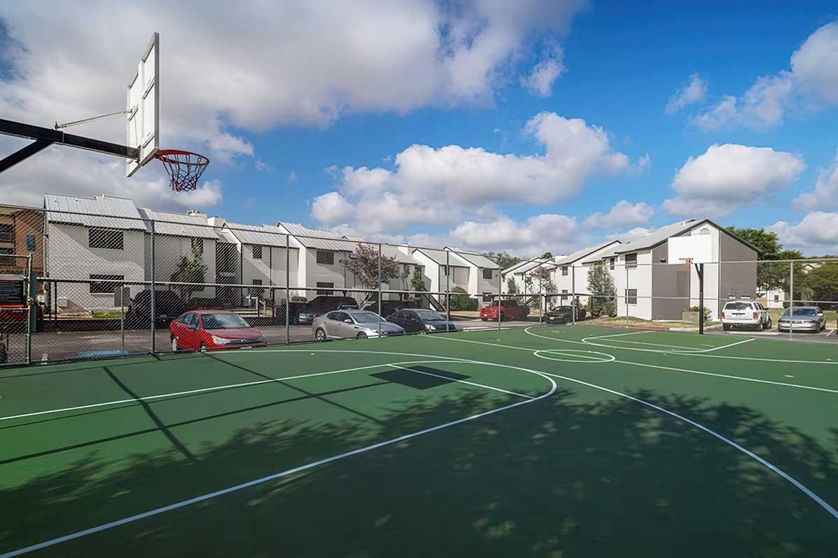 A basketball court with a hoop in the foreground, surrounded by a chain-link fence. In the background, there are residential buildings and parked cars under a blue sky with fluffy white clouds. The court is freshly painted with a vibrant green surface.