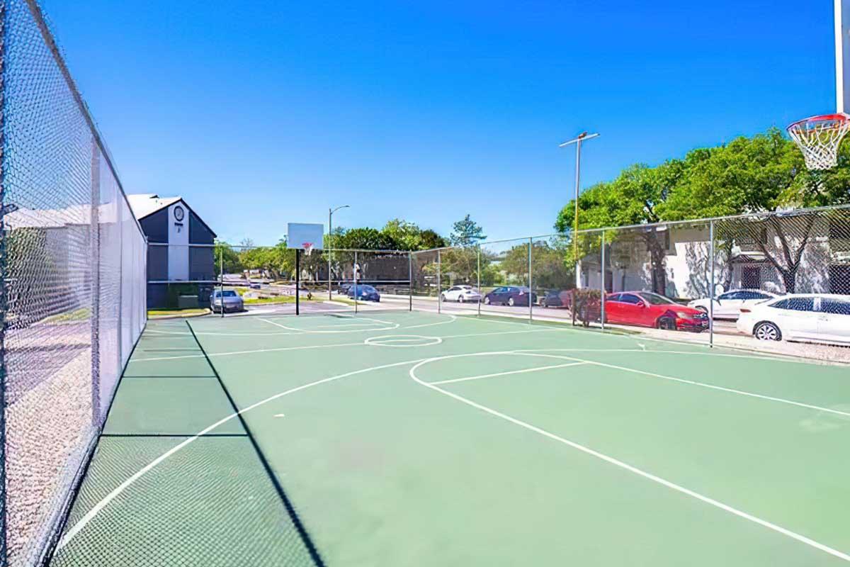 A wide view of an empty basketball court enclosed by a chain-link fence, featuring clear blue skies and a few trees in the background. Nearby, several parked cars are visible along the street, and there are some residential buildings in the distance.