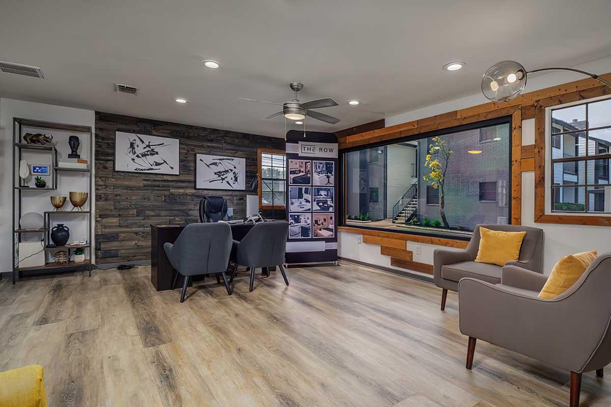 A modern office space featuring wooden accents, a large window with a view, and stylish furniture. There are two gray armchairs, a desk with a computer, and decorative items on shelves. Natural light illuminates the room, creating a welcoming atmosphere.